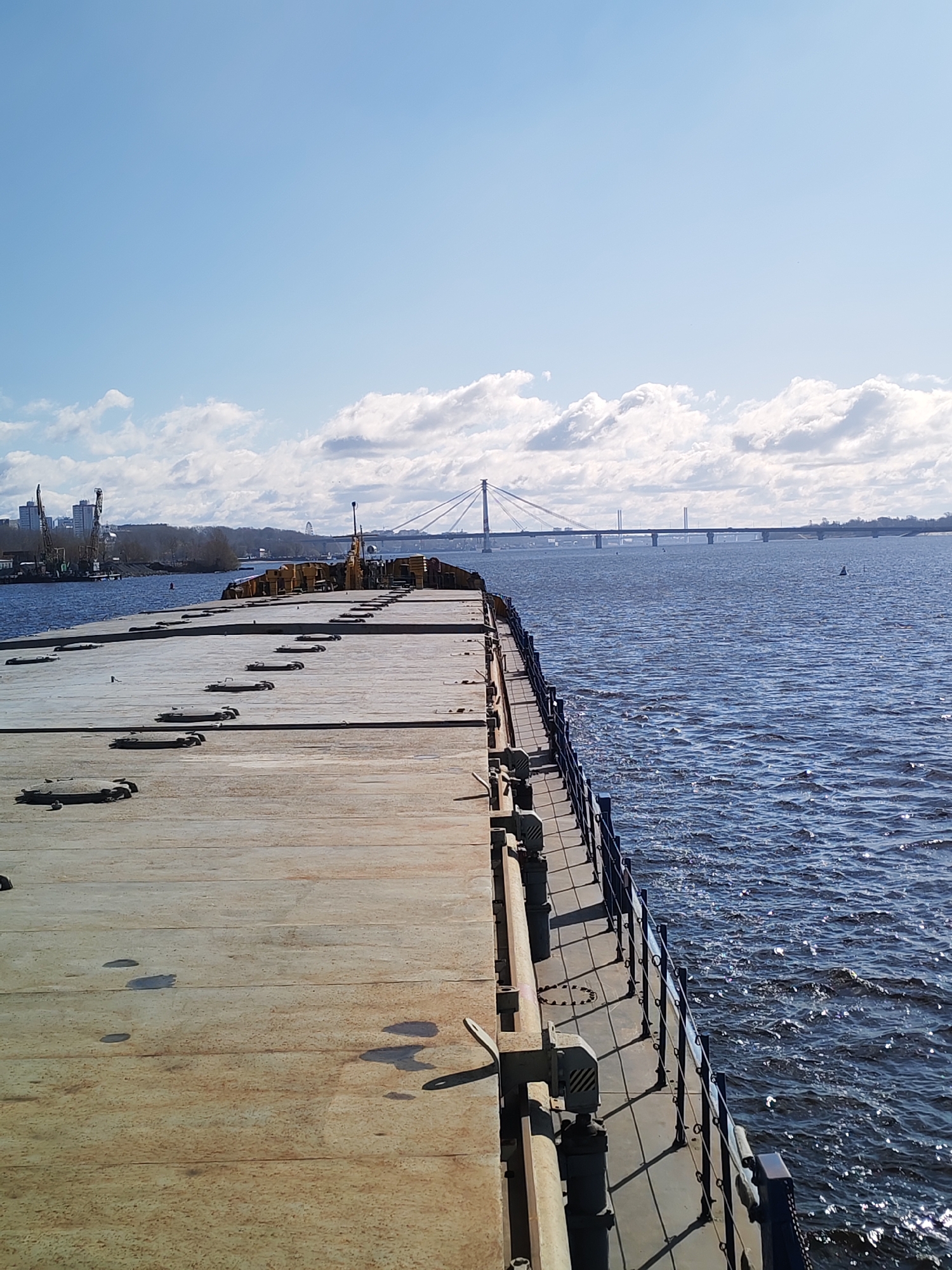 The image displays an expansive outdoor scene, dominated by a large, flat-topped structure, likely a barge or a wide pier, in the foreground, stretching from the bottom left towards the distant horizon. Its surface is a weathered, light-colored material, possibly concrete or wood, embedded with numerous circular and rectangular metal hatches. Along the right edge of this structure, a dark-colored railing with vertical posts and horizontal ropes or chains runs parallel to the water.
To the right, a wide body of dark blue water, possibly a river or a lake, ripples under the bright sunlight, creating shimmering reflections. Small buoys or markers are visible on the water's surface in the mid-ground. In the distance, a prominent cable-stayed bridge spans the water, characterized by tall support towers and a web of cables. Behind it, another, simpler bridge with multiple piers extends across the water. On the far left, a distant city skyline is visible, featuring various buildings and what appear to be industrial cranes or structures along the water's edge. A Ferris wheel is also discernible on the distant bank.
The sky is a clear, vibrant blue, dotted with scattered, fluffy white cumulus clouds. The scene is brightly lit, indicating it is daytime, likely late morning or early afternoon. The overall impression is one of calm and openness. There are no people visible, nor any specific activities taking place. No discernible text is present in the image.