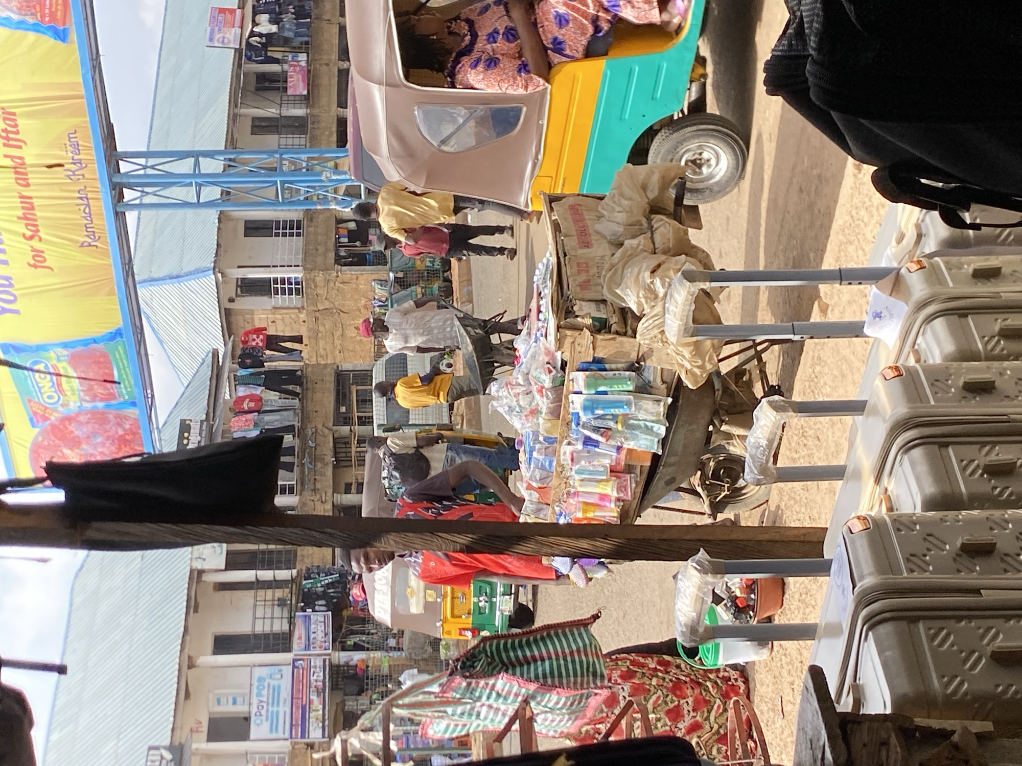The image shows a street scene in Bukuru, Nigeria. The buildings are multistory, with shops on the ground floor. There are people walking in the street, and a three-wheeled vehicle is also visible. The street is crowded, with goods displayed for sale. There are a number of objects, including luggage, packaged goods, and bags. A blue banner, “for Sahur and Iftar”, is displayed, indicating that it may be the month of Ramadan.