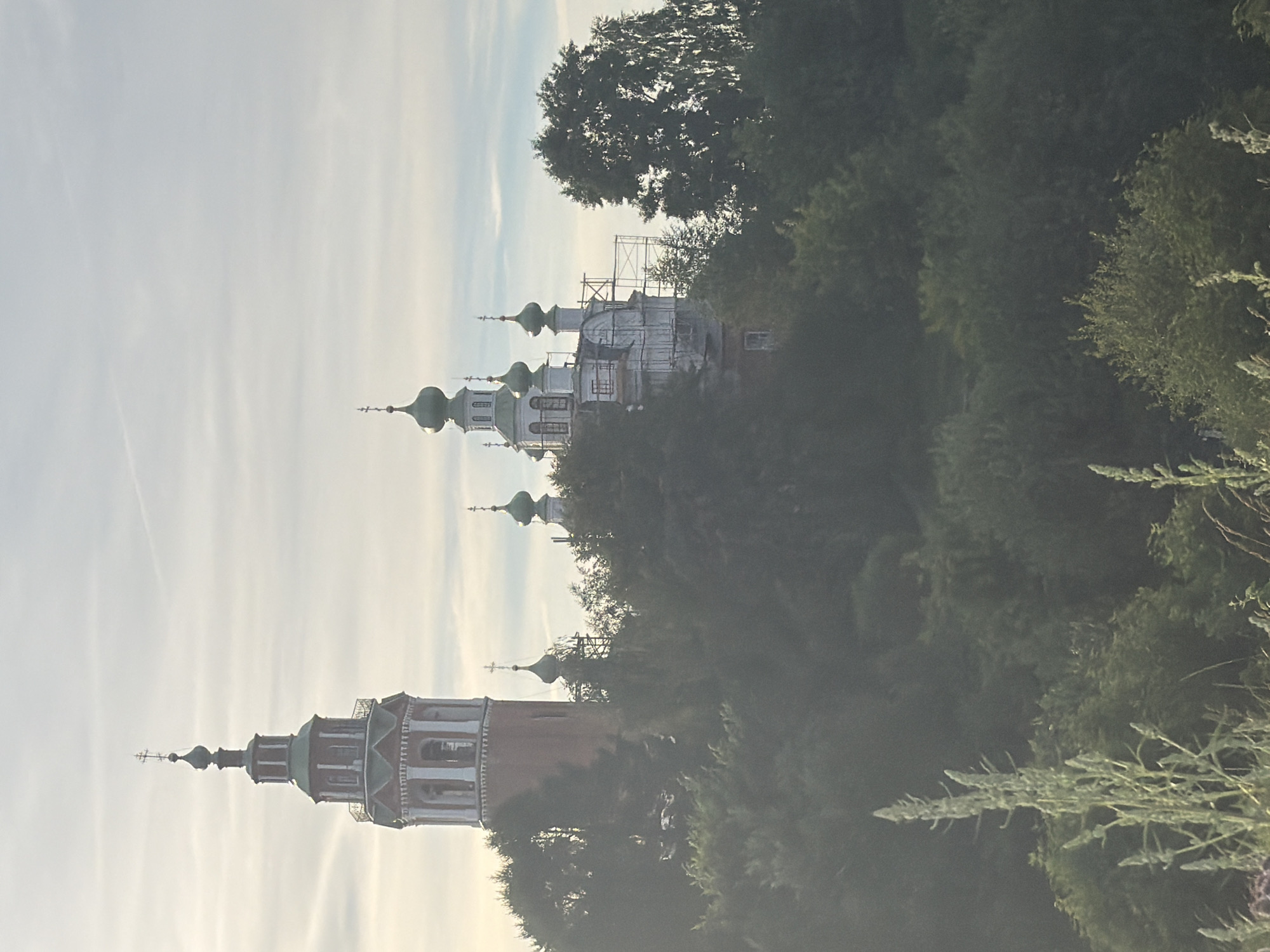 This image shows the bell tower and domes of the Church of the Assumption of the Blessed Virgin Mary in Cherdyn, Russia. The bell tower is tall and red with a green roof and a cross at the top. The domes of the church are green and bulbous, and some of them are surrounded by scaffolding, suggesting renovation work. The buildings are situated on a wooded hill, with lush green trees and foliage surrounding them. In the foreground, there are tall, pale green plants. The sky is overcast with thin, wispy clouds.
