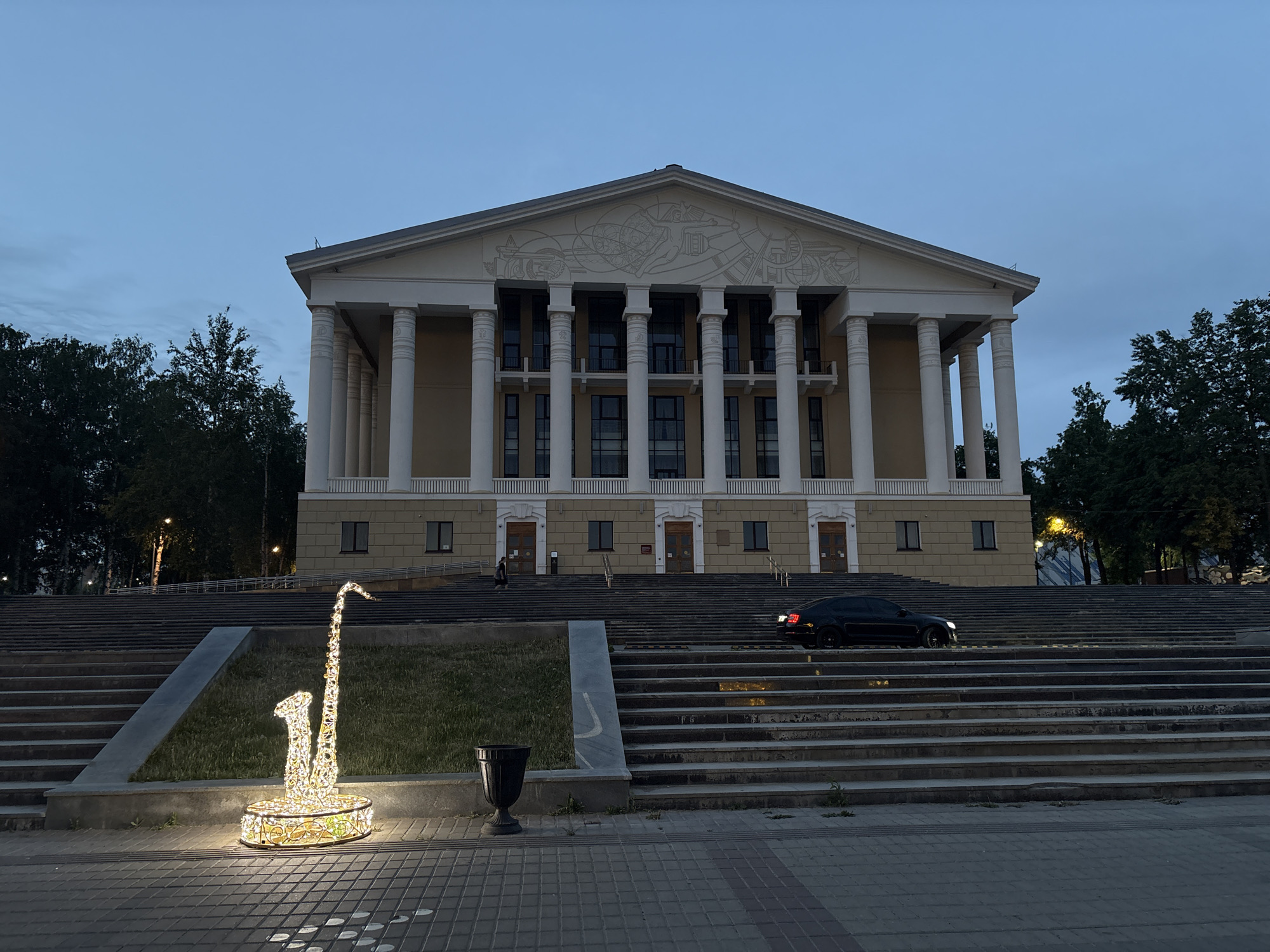 The image depicts the exterior of the Palace of Culture in Votkinsk, Russia, at dusk. The building is a large, classical structure with a portico supported by tall white columns and a pediment adorned with a relief sculpture. A broad set of stairs leads up to the entrance. In the foreground, a decorative light sculpture resembling a saxophone stands on the paved plaza. A black car is on the staircase leading to the entrance. Trees are visible on both sides of the building. A few individuals appear on the stairs near the entrance.