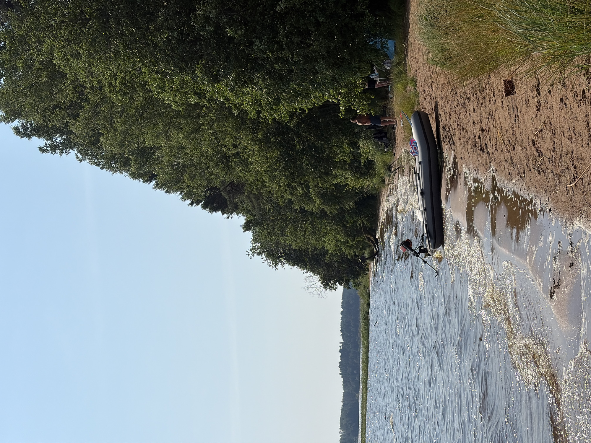 A medium shot captures a sunny day at a sandy lake shore in Vaskelovo, Russia. The scene is dominated by a tranquil lake on the left, with small waves lapping at the shore, reflecting the clear blue sky. A line of lush green trees forms a dense backdrop along the right side of the image, extending towards the horizon.
On the sandy beach, an inflatable boat with an outboard motor is partially beached. A few people are visible near the trees. One man stands shirtless in blue shorts, facing away from the camera. Further back, two figures, one appearing to be a woman in dark clothing and another person, are near a table. Camping equipment, including chairs and a blue tent, is scattered on the sand. Fishing rods are propped up near the water's edge, suggesting recreational activities. The overall atmosphere is relaxed and typical of a summer outing at a lakeside location.