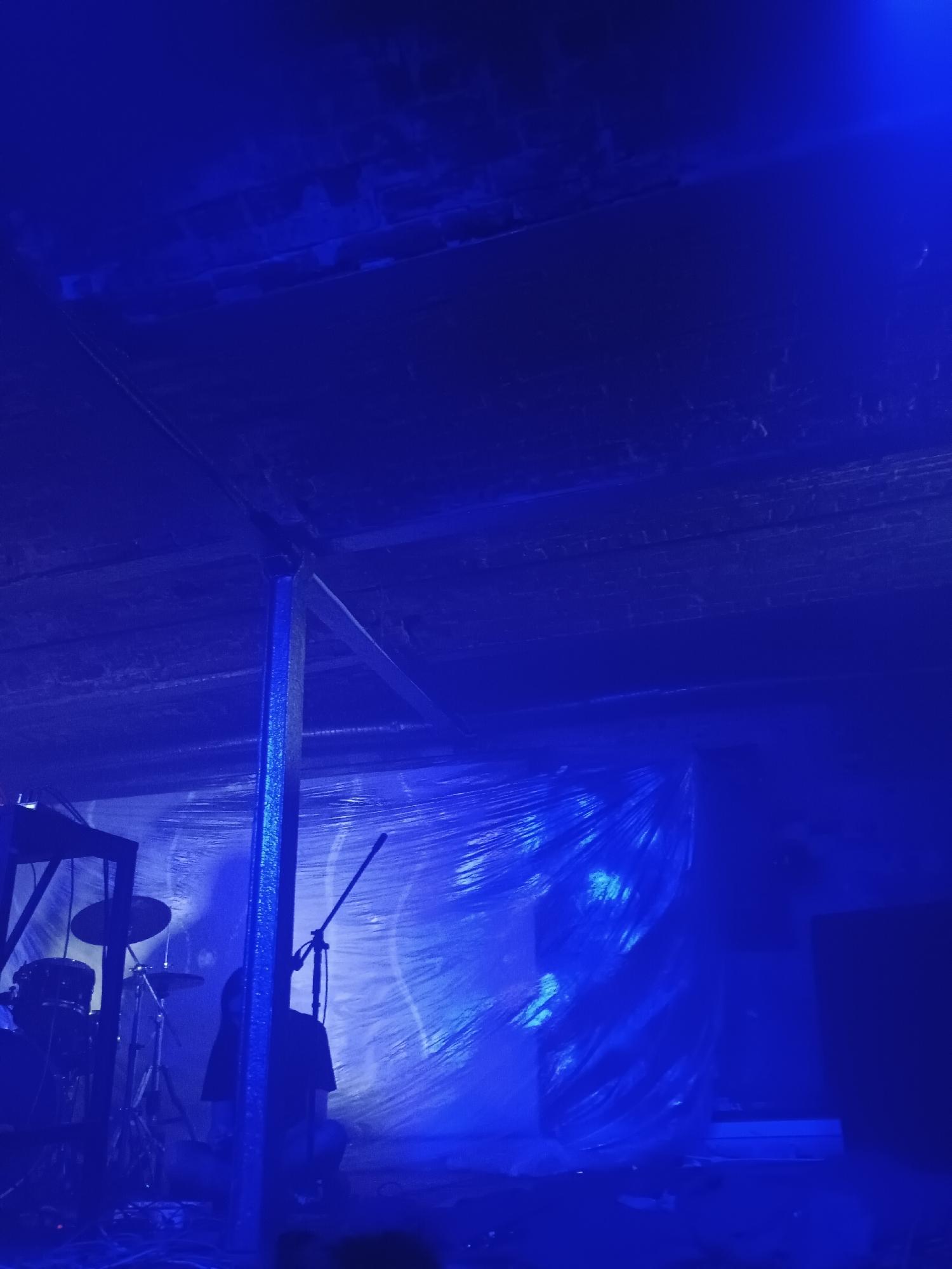 The image depicts an indoor scene illuminated by a strong blue light, suggesting a stage or performance area. In the foreground left, a drum set is visible, consisting of drums and cymbals. To the right of the drum set, a person wearing a dark shirt stands behind a microphone stand. The background features a wall covered in what appears to be plastic sheeting, creating a textured and reflective surface. The ceiling, made of exposed beams and wooden planks, is also visible. There are no discernible elements that indicate a specific location within Moscow, Russia.
