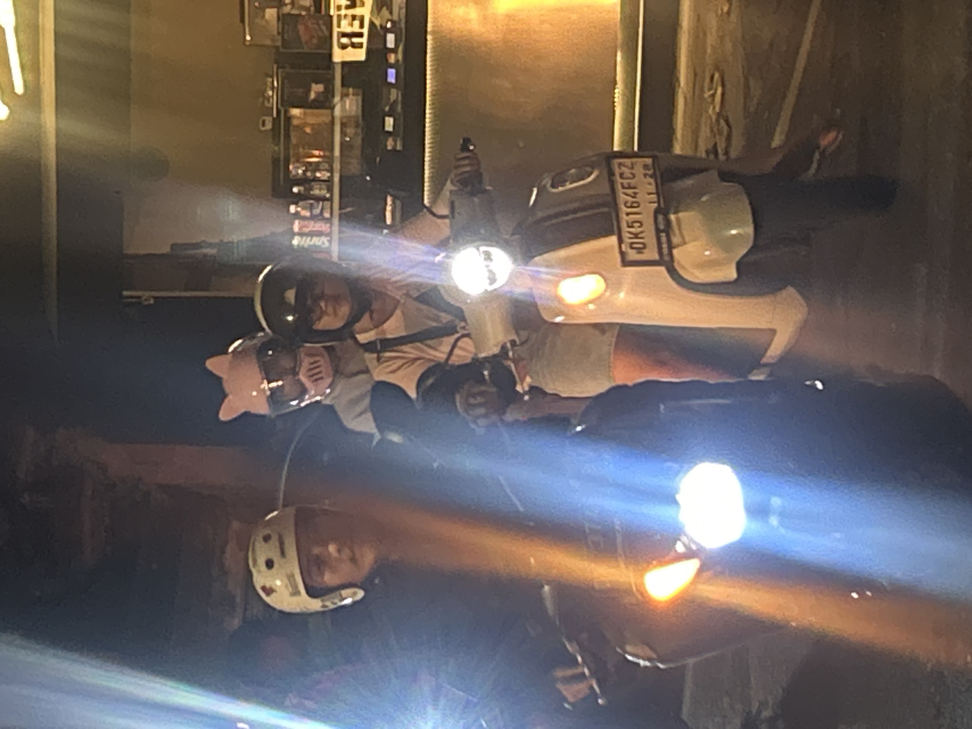 In North Kuta, Indonesia, three individuals are seen riding on a scooter at night. The person in the front, wearing a white helmet with decorative markings, appears to be the driver. Behind them, a person in a pink helmet with cat ears is seated, and another person is positioned behind that individual. The scooter has a license plate that reads "DK5164FCZ 11-28". The environment suggests an urban setting with illuminated signage and building facades visible in the background. The lighting conditions are low, with artificial lights from the scooter and surrounding areas contributing to the scene.