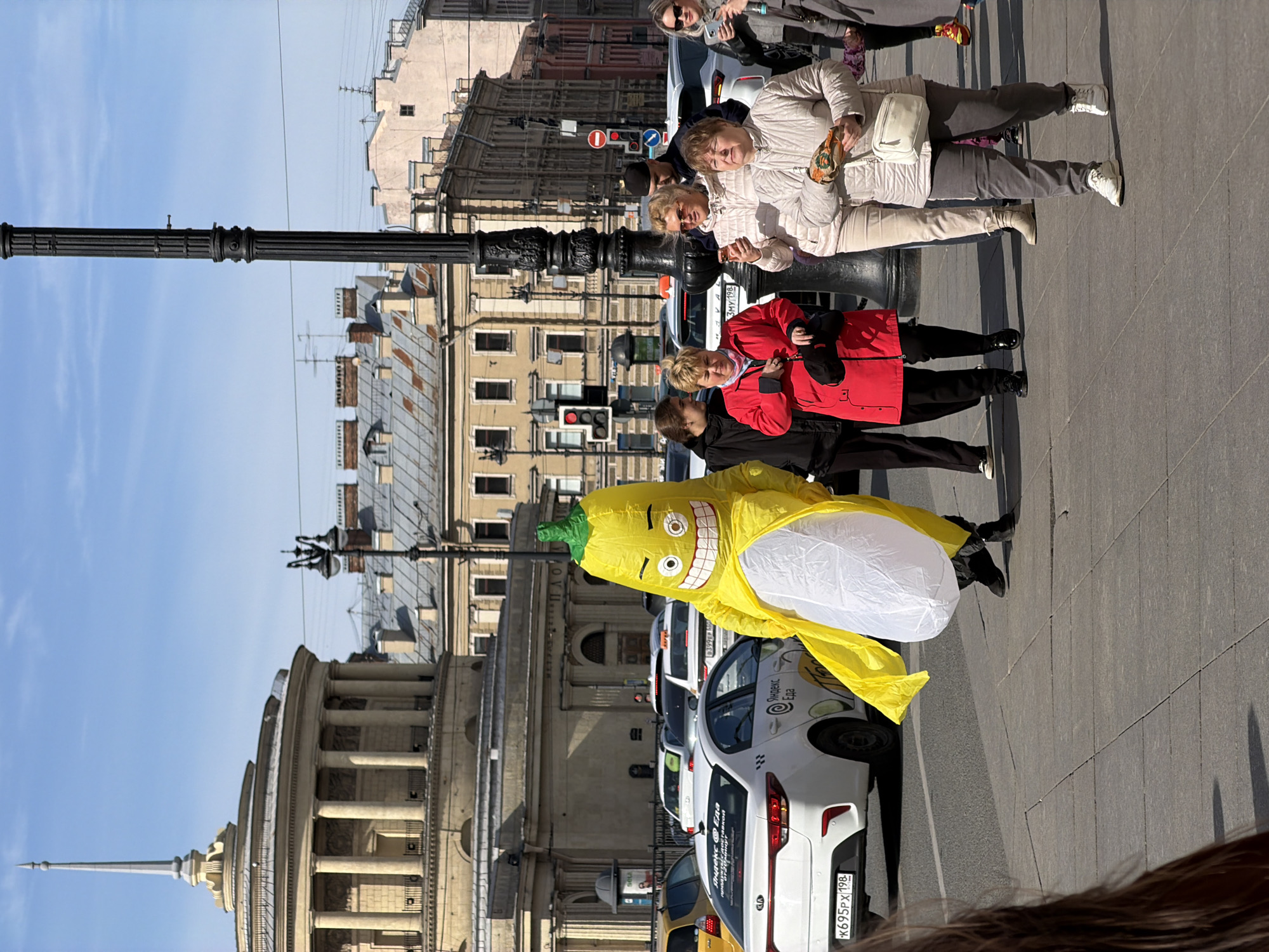 The image shows a street scene in Saint Petersburg, Russia, on a sunny day. The background includes a view of Nevsky Prospekt with various buildings. A distinctive building on the left features a tall spire. Several cars are parked along the street, with a white taxi prominently displaying visible Cyrillic text. A person in a yellow inflatable banana costume is walking down the sidewalk. Several individuals are standing on the sidewalk. A black lamppost is also present.