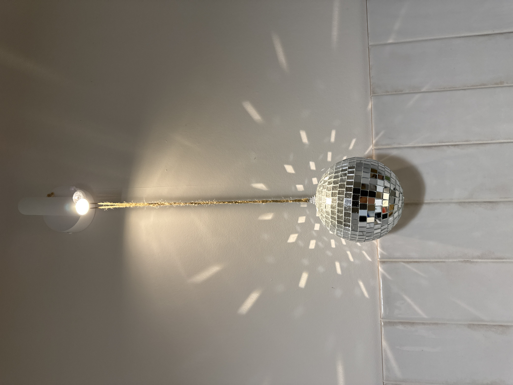 The image shows a disco ball hanging from a wall light fixture. The disco ball is silver and reflects light onto the wall and a section of horizontally laid white tiles. The light fixture is white and cylindrical, extending from the wall. A length of twine connects the light fixture to the disco ball. The location is unknown, and there is no evidence it is located in Izhevsk, Russia. There are no people visible in the image.