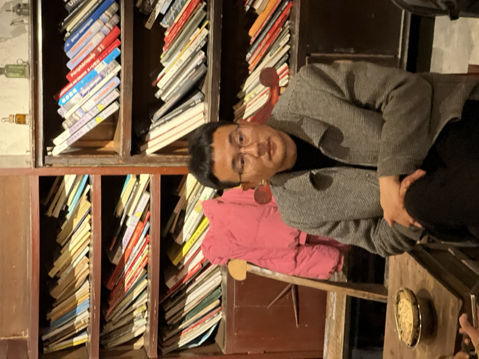 A man in glasses and a gray jacket sits in front of a bookshelf filled with books. He is turned slightly to the right, looking at the camera. His right hand rests on his left knee. A pink jacket hangs on the chair. On the table to his right is a small bowl filled with a light-colored substance. Part of another person's hand is visible on the far right edge of the image. The bookshelves are full and the books are tilted.