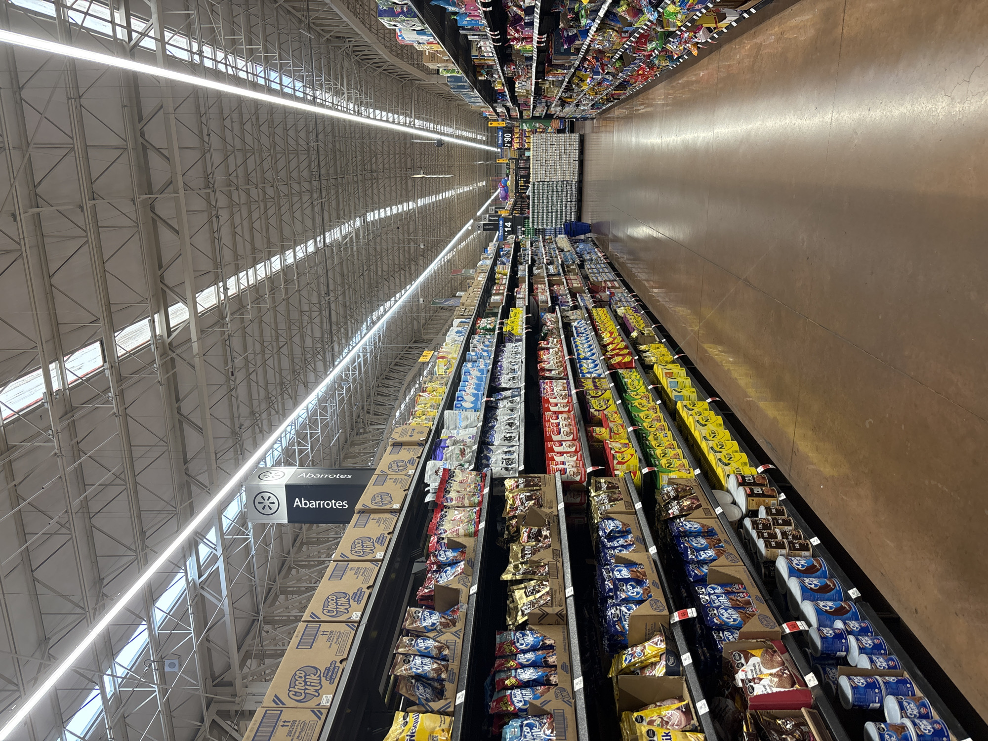 Aisles of a supermarket are seen in this eye-level shot. The shelves are filled with various items, the metal ceiling structure is visible, and the floor is smooth and reflective. A sign labeled "Abarrotes" hangs from the ceiling. The location is a supermarket in San Bernardino Tlaxcalancingo, Mexico.