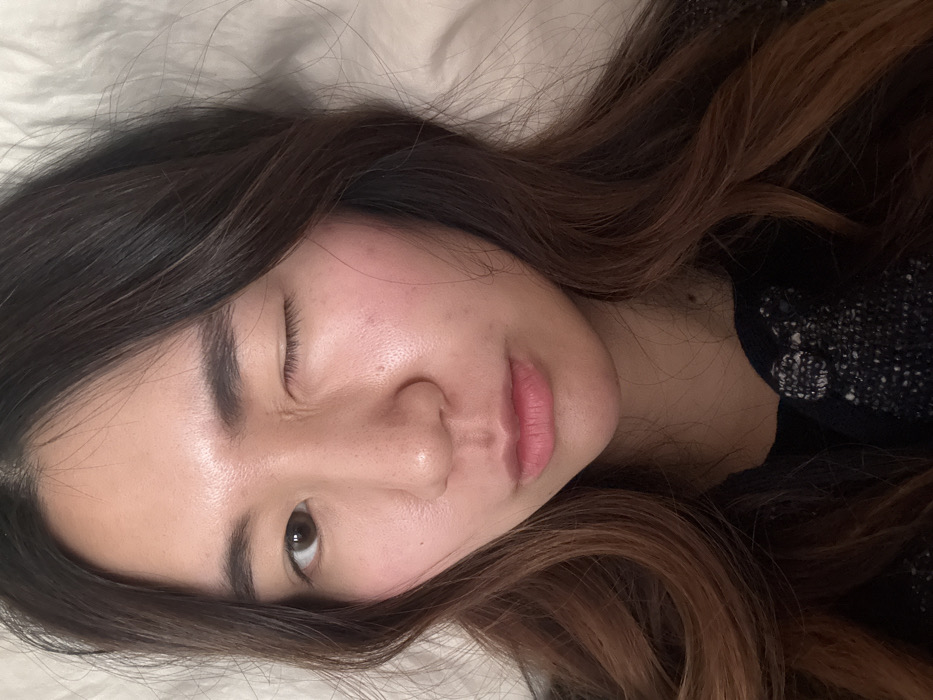 The image displays a close-up portrait of an adult female. The specific location within Paris, France, cannot be determined from the visual evidence, as the background is an out-of-focus, light-colored, plain surface, consistent with an indoor setting lacking identifiable features.
The subject has long, dark brown hair with lighter, possibly auburn, highlights, styled in loose waves that frame her face and extend over her shoulders. Her complexion is light to medium, with a visible slight flush on her cheeks.
Her right eye (viewer's left) is open, revealing a brown iris. Her left eye (viewer's right) is tightly closed in a wink, creating fine wrinkles around the orbital area. Her eyebrows are dark and appear relatively thick. The subject's nose is centered, and minor skin pores are discernible. A small reddish mark or blemish is present on the philtrum, directly above her upper lip. Her lips are closed, slightly pursed, and exhibit a natural pink hue.
She is wearing a dark garment, potentially black, with a textured or speckled pattern visible on the upper chest and shoulder area, suggesting a knit or woven material with lighter flecks or embellishments. The lighting is soft and even, illuminating her face directly.