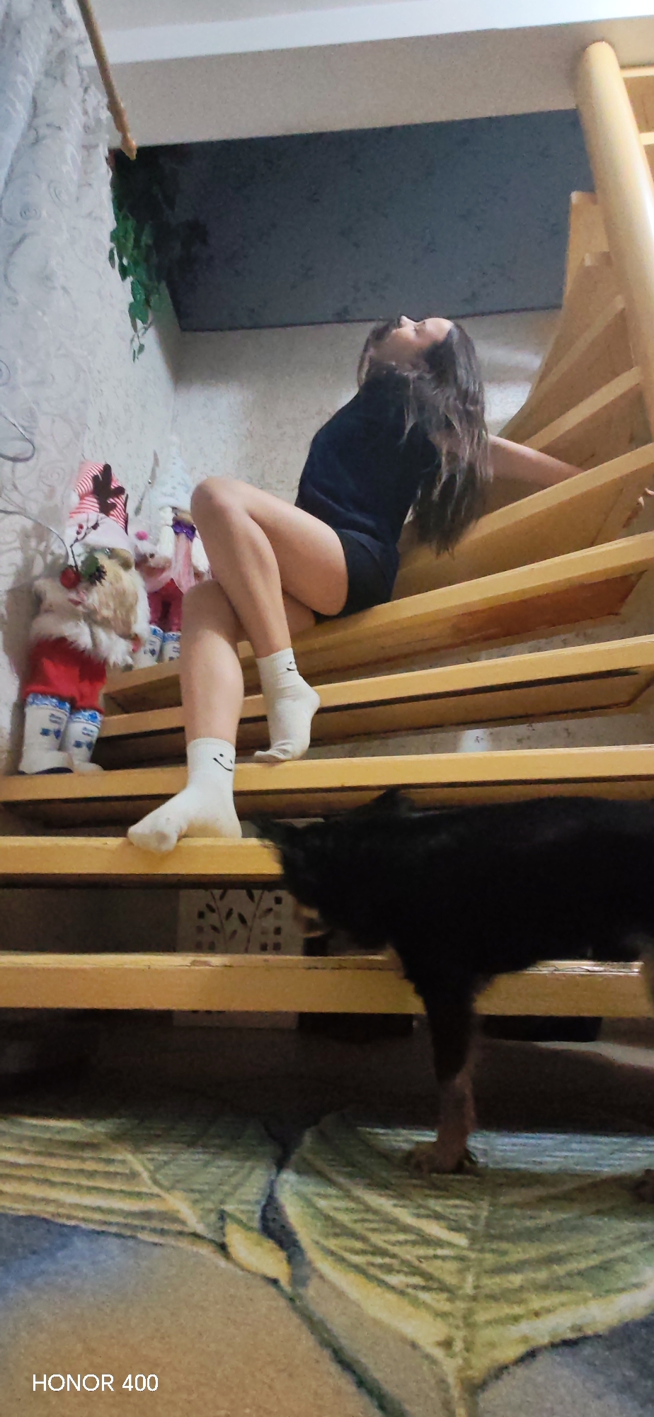 A woman is sitting on wooden stairs inside an unidentified building in Noviy, Russia. She is wearing a dark shirt and shorts, and white socks with smiley faces on them. To her left, on a higher step, are three decorative gnome figures. A black dog is partially visible on a lower step. The floor is covered with a rug with a leafy pattern. The wall on the left is textured and has some greenery. The staircase has a light wood finish and a vertical support post. The background includes a white wall and a dark section above it. The photo was taken with a Honor 400 camera.