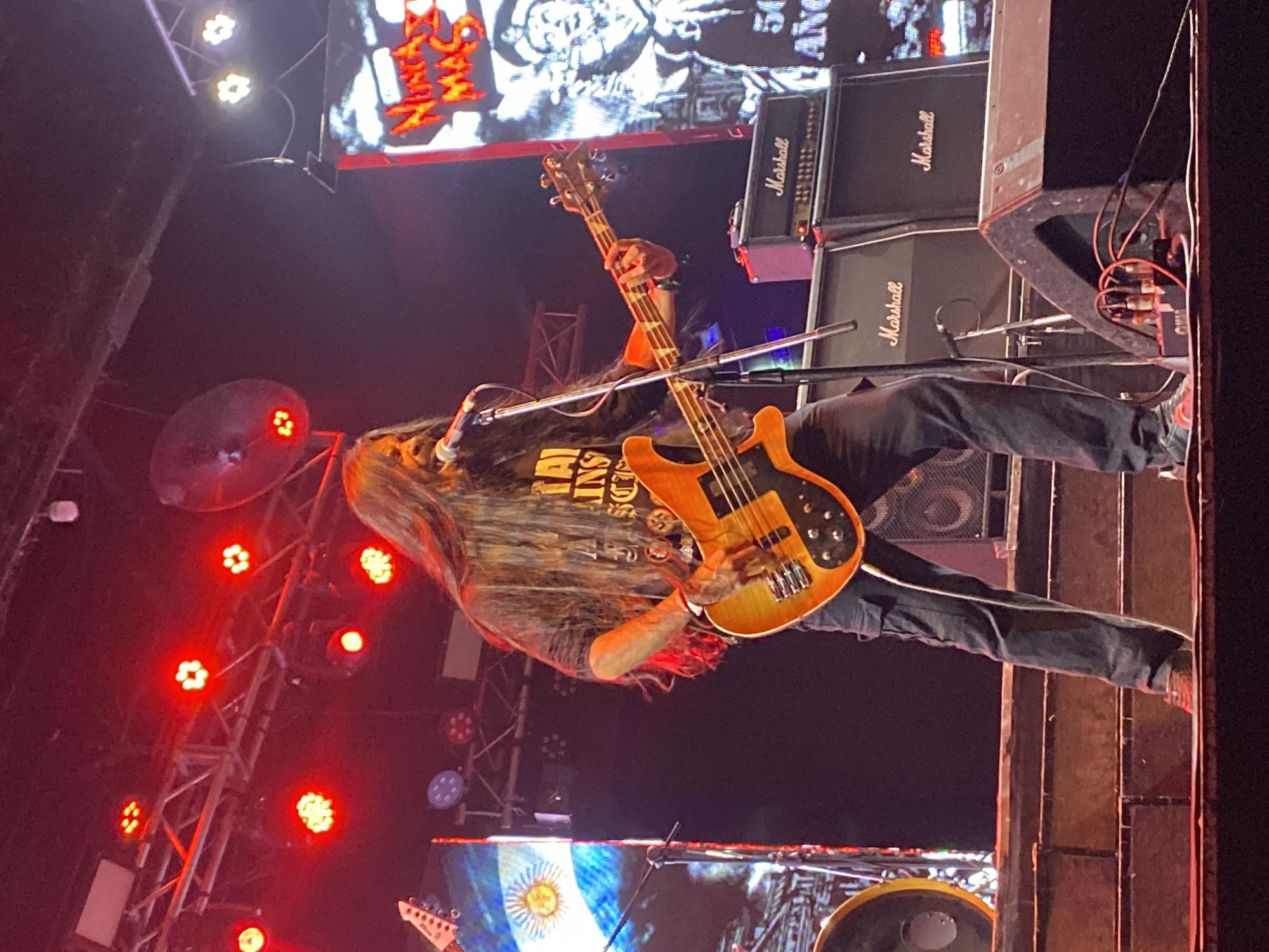A male musician with long, dark hair is performing on an illuminated stage within a dark venue. He stands centrally, wearing a dark t-shirt with light lettering and dark cargo pants, playing a four-string, sunburst-finish bass guitar with a black pickguard. His left hand is positioned on the fretboard, and his right hand plucks the strings. He sings into a silver microphone affixed to a stand.
The stage setup includes multiple Marshall amplifier heads and speaker cabinets stacked on the right side, along with a floor monitor in the foreground. A drum kit, including a bass drum with a similar finish to the bass guitar, is partially visible on the left. Overhead trussing supports numerous red LED stage lights and two circular industrial fans.
Two large LED screens form the backdrop: the left screen displays the Argentinian flag with the "Sun of May" emblem. The right screen features prominent yellow and red text "NUNCA MAS" at the top, above a black-and-white graphic of a uniformed figure (possibly military or naval) pointing. Partially visible text "50 AÑOS" appears below this figure. The stage is multi-tiered, with visible steps.
The presence of the Argentinian flag and the "NUNCA MAS" slogan indicates the event is located in Argentina, specifically within the city of Buenos Aires. No further precise venue details are identifiable from the image.