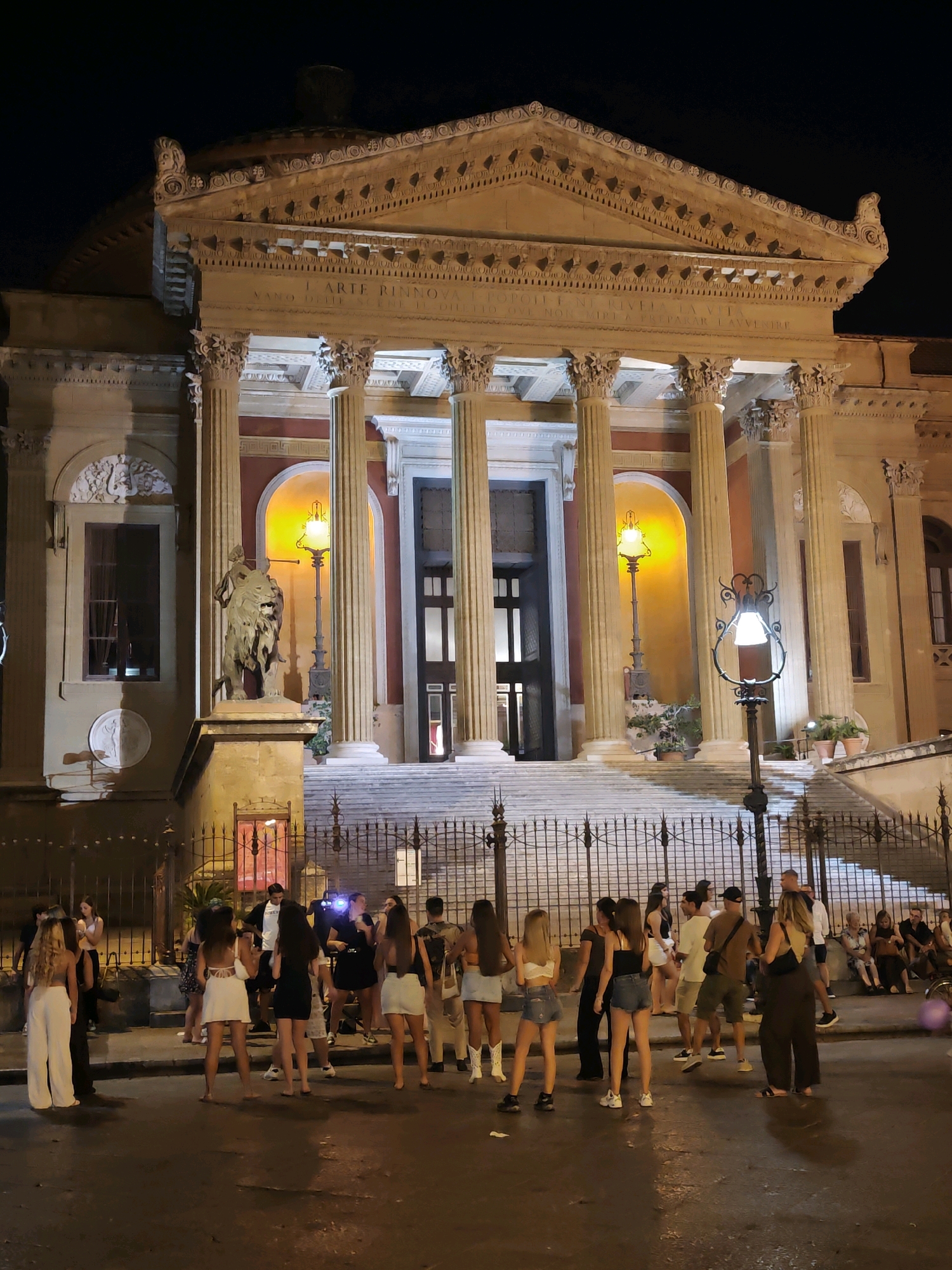 The image captures a nighttime exterior scene of the Teatro Massimo in Palermo, Italy. The grand opera house features a classical facade with a pediment, Corinthian columns, and arched entrances, all illuminated by warm yellow lights. In front of the building, a statue of a man standing over a lion is visible on a pedestal. A wrought-iron fence separates the sidewalk from the building's grand staircase.
On the street in front of the theater, a group of young people are gathered. They appear to be dressed for a night out, with women in dresses, skirts, and sandals, and men in casual attire. Some are standing and talking, while others are seated on the steps of the theater or the surrounding low walls. The street is wet, reflecting the lights from the theater and street lamps. The overall atmosphere suggests a lively evening outside a prominent cultural landmark.
