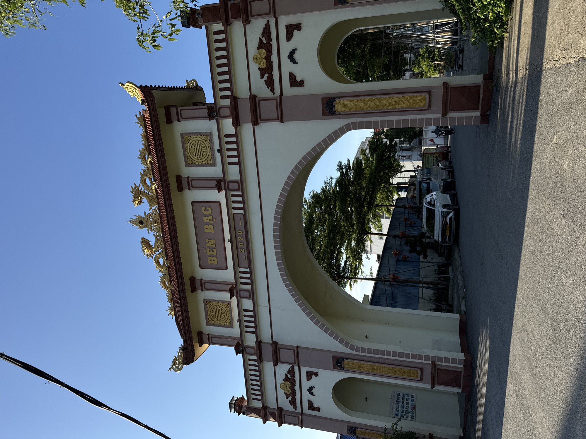 The image shows an ornate gateway in Hà Nội, Vietnam. The structure, painted in cream with reddish-brown accents and gold embellishments, features a central arch and smaller side arches. The words "BẾN BẮC" and "2020" are displayed on the gateway. Intricate dragon details adorn the top of the structure. The gateway frames a view of a street scene with parked cars, trees, and buildings. The sky above is clear and blue. A power line is visible in the lower left corner. The street pavement is visible in the foreground.