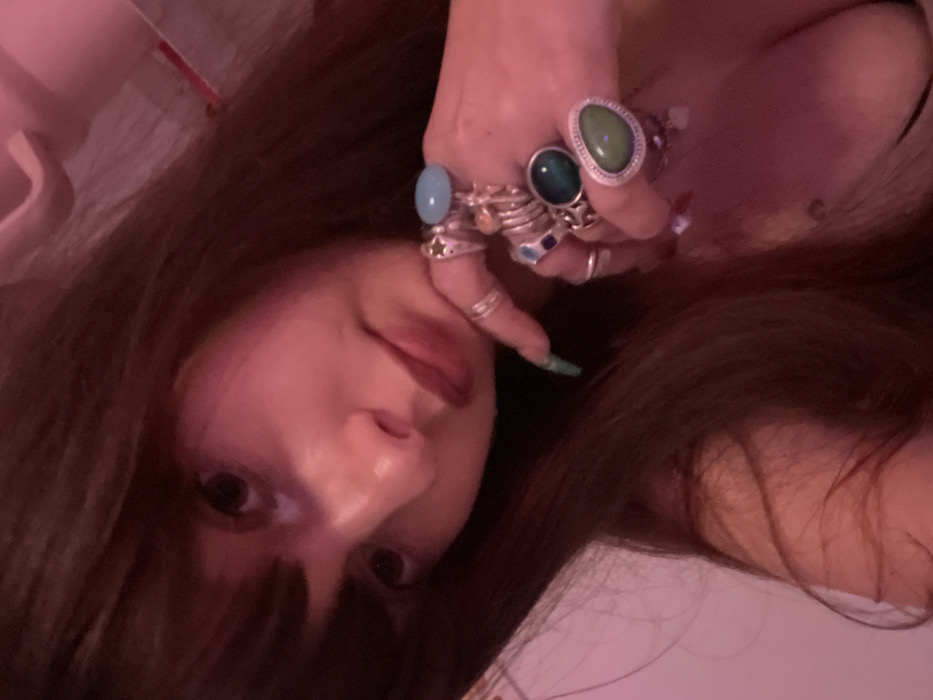 A woman with long brown hair and bangs poses with her face close to the camera. She has fair skin, brown eyes, and red lipstick. Her left hand is raised, adorned with multiple rings of varying sizes and designs, including stones in shades of blue, green, and silver. The image is close-up, and the lighting is soft and warm, giving the scene a romantic and intimate feel.