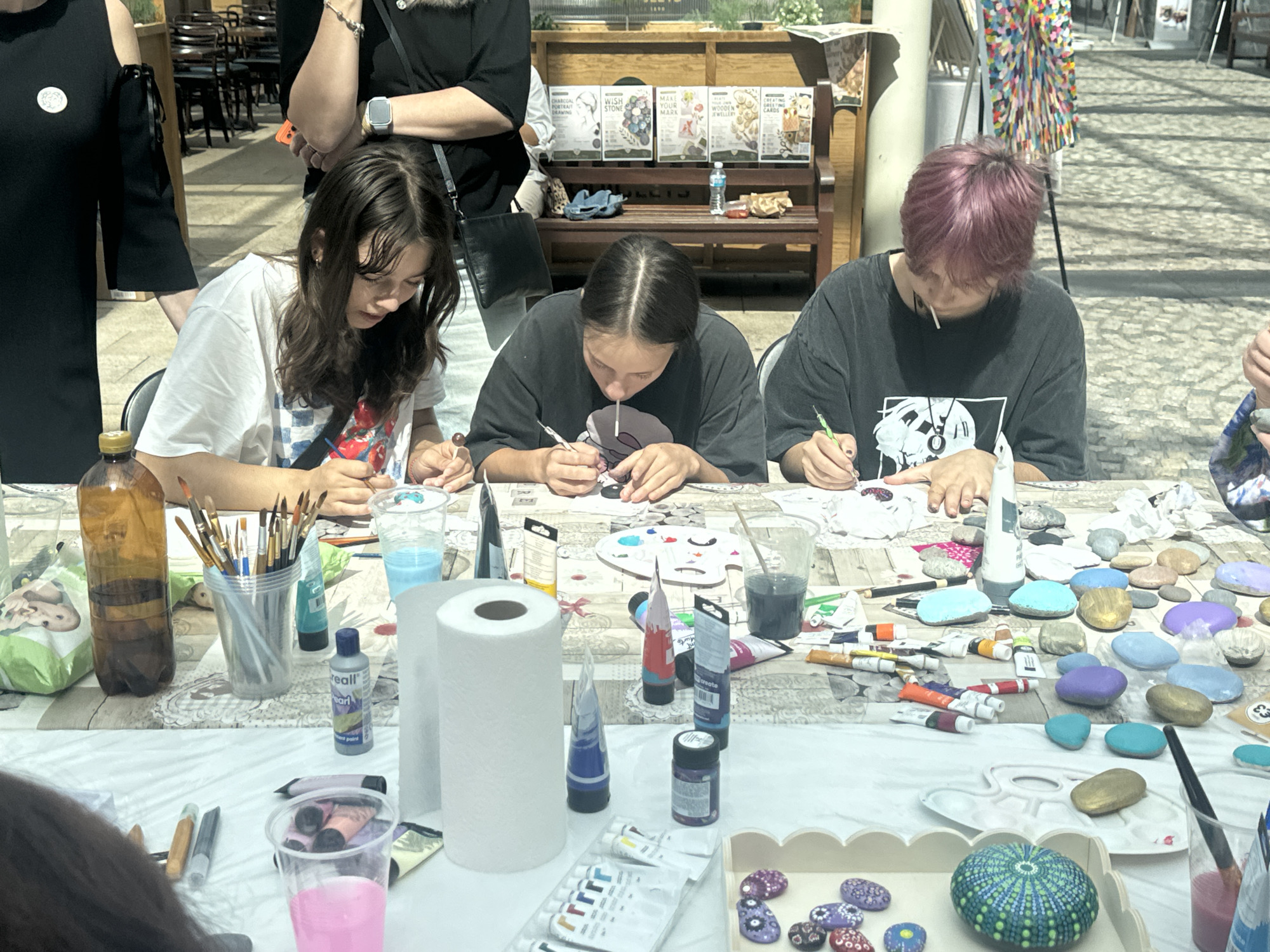 The image depicts a group of three individuals, two young women and one person with pink hair, engaged in painting small stones at an outdoor market stall. They are seated at a table covered with a white tablecloth, which is laden with art supplies including paint tubes, brushes, water cups, and a roll of paper towels. The individuals are focused on their work, with some using fine brushes to paint intricate designs on the stones. In the background, other market stalls, seating areas, and a painting on an easel are visible, suggesting a vibrant, public setting. The overall scene suggests a creative workshop or a craft activity taking place in a community space. The presence of Irish signage in the background and the distinct style of the market stalls indicate the location is in Kilkenny, Ireland.