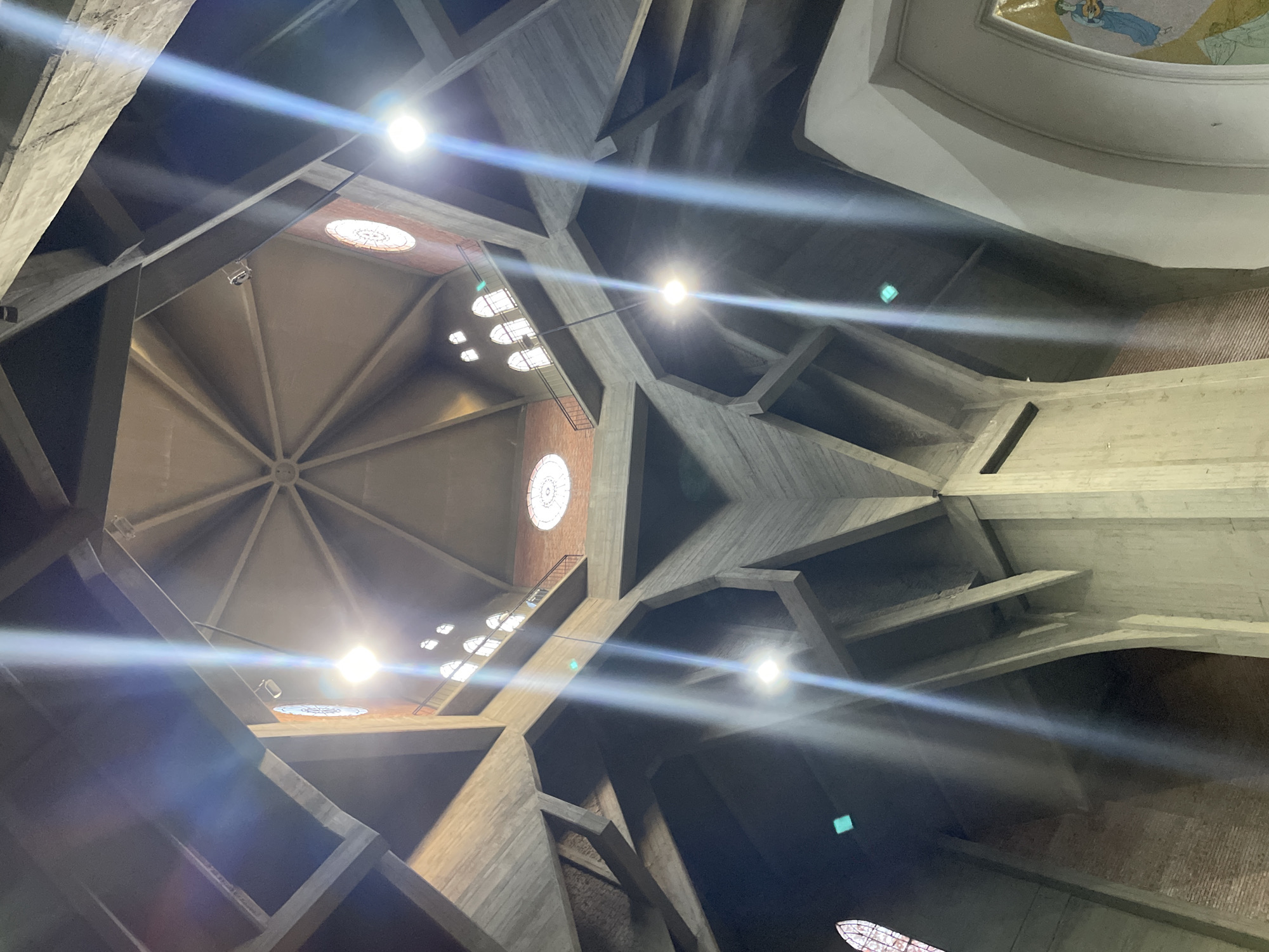 The image captures an upward view of a striking interior architectural space, likely a large church or cathedral, located in Vicente López, Argentina. The dominant features are massive, angular concrete beams and structural elements that form a complex, geometric ceiling. These concrete structures are unadorned, reflecting a brutalist or modernist aesthetic.
Bright, artificial lights with pronounced lens flares illuminate the scene, creating a sense of dramatic lighting and emphasizing the interplay of light and shadow on the textured concrete surfaces. Several windows are visible, including circular stained-glass windows with intricate patterns and elongated, possibly rectangular, windows or skylights that also appear to be stained or patterned.
In the upper right portion of the frame, a curved, white ceiling section features a faint mosaic or fresco depicting human figures, further suggesting a religious context. Towards the left, a set of dark stairs or a walkway structure can be seen leading upwards along one of the concrete walls, indicating multiple levels within the building. A small, white security camera or speaker is mounted on one of the beams. The overall impression is one of grandeur and architectural innovation, with a focus on form, material, and light. No discernible text is visible in the image.