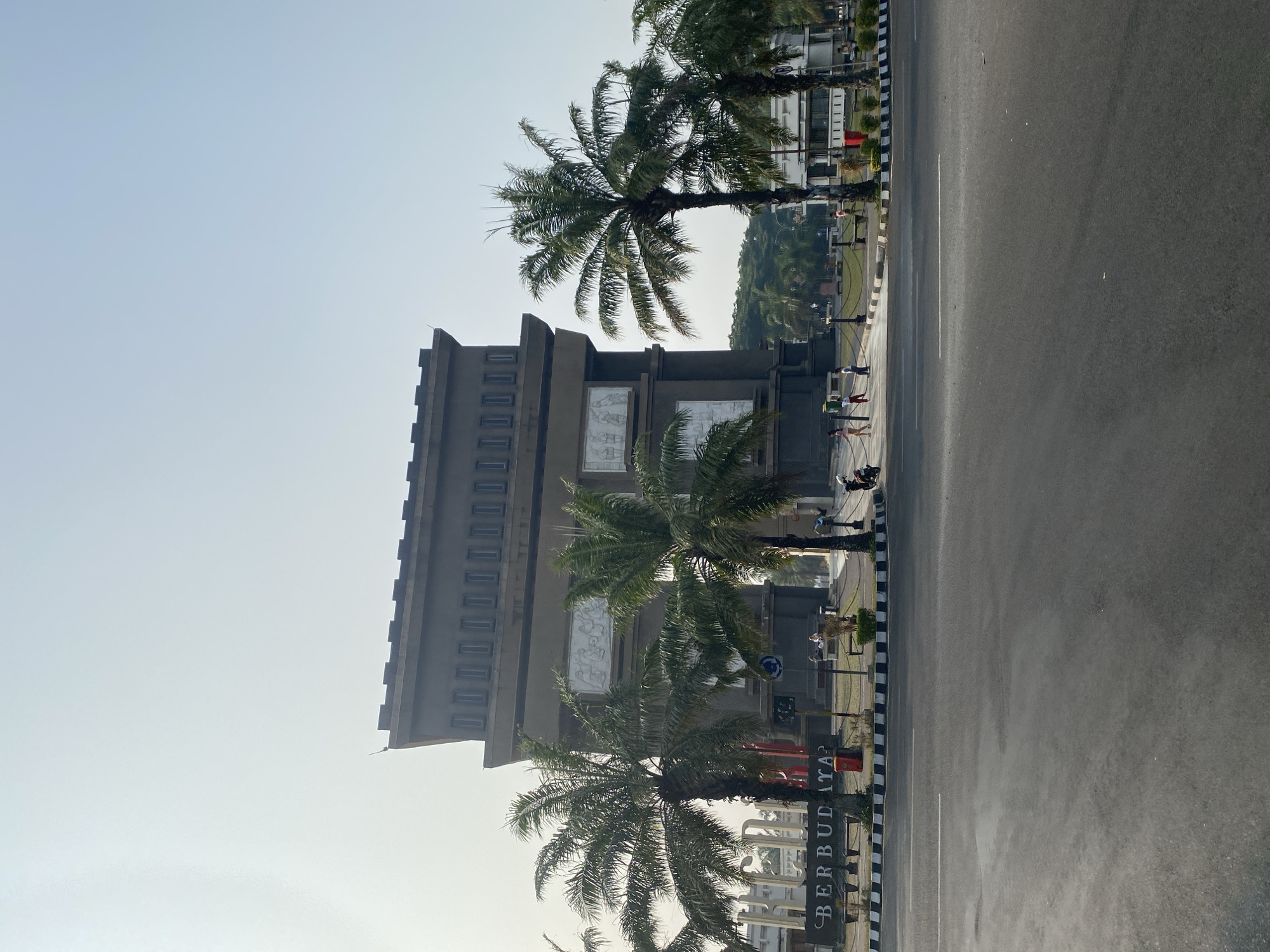 This image captures the Tugu Yogyakarta landmark in the city of Ngasem, Indonesia, on a bright, clear day. The monument is a large, square structure with a crenellated top, resembling a fortress. Its facade is adorned with relief sculptures. In front of the monument, several palm trees stand tall, their fronds fanning out against the sky. A street with a white line marking the lane runs across the foreground. On the median in front of the monument, the word "BERBUDAYA" is spelled out in large letters. A few people are visible in the midground, some standing and others walking, near the monument's base. One person is on a motorcycle to the right of the frame. The environment appears to be a public space, possibly a park or plaza surrounding the monument, with some greenery and benches visible. The overall impression is one of a significant cultural site in an urban setting.