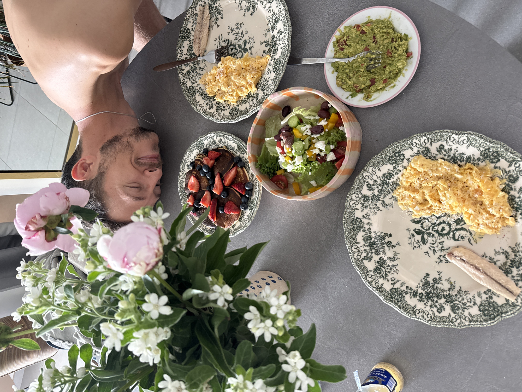 A man with closed eyes and a silver necklace is visible in the top left of the image, turned upside down. The frame also shows a table featuring breakfast items. There are plates with scrambled eggs, a plate of guacamole, chocolate pancakes with berries, and a salad. A vase of flowers is on the table as well. There is no information indicating the location of this scene is in Tel-Aviv, Israel.