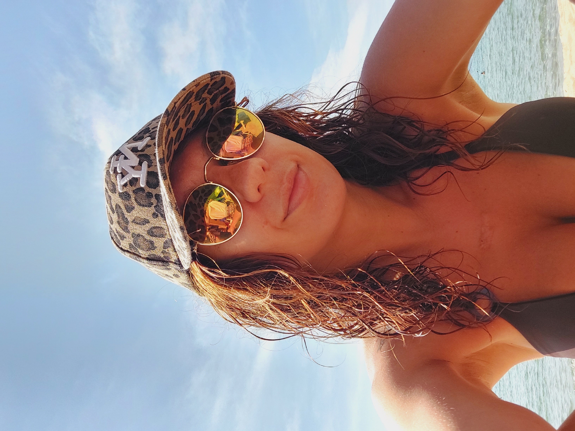 A woman with wet, wavy auburn hair is photographed from the chest up, outdoors, under a bright blue sky with sparse white clouds. She is wearing a leopard-print baseball cap with a white "YM" logo on the front and round, reflective sunglasses. Her shoulders and upper chest are visible, revealing a dark bikini top. The background shows a calm, light blue sea and a sandy beach. The overall impression is one of a casual, sunny day at the coast. Given the context of Sozopol, Bulgaria, this scene likely depicts a tourist or local enjoying a beach on a warm day.