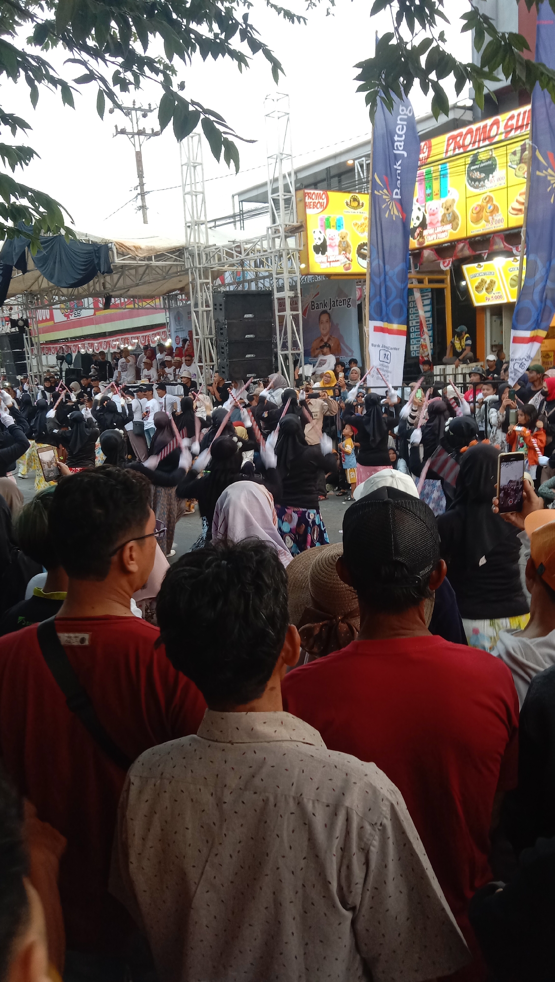 This image captures an outdoor event, likely a festival or performance, in Jatisawit, Indonesia. In the foreground, a crowd of people is gathered, with their backs to the camera, observing the activity on a stage in the background. The stage is equipped with large speakers and a metal truss structure, suggesting a professional setup.
A group of performers, predominantly women wearing headscarves, are actively engaged in what appears to be a choreographed dance on the stage. They are holding thin, rod-like objects with what seem to be flags or fabric attached, which they are waving. The performers are dressed in a mix of black and colorful patterned attire.
Behind the stage, there are buildings with banners and signs, including one that reads "PROMO HEBOH" and another that says "Bank Jateng," indicating commercial sponsorship for the event. The overall atmosphere suggests a lively community gathering. The background also shows trees and overhead power lines, typical of an urban or semi-urban environment. The lighting indicates it is daytime.