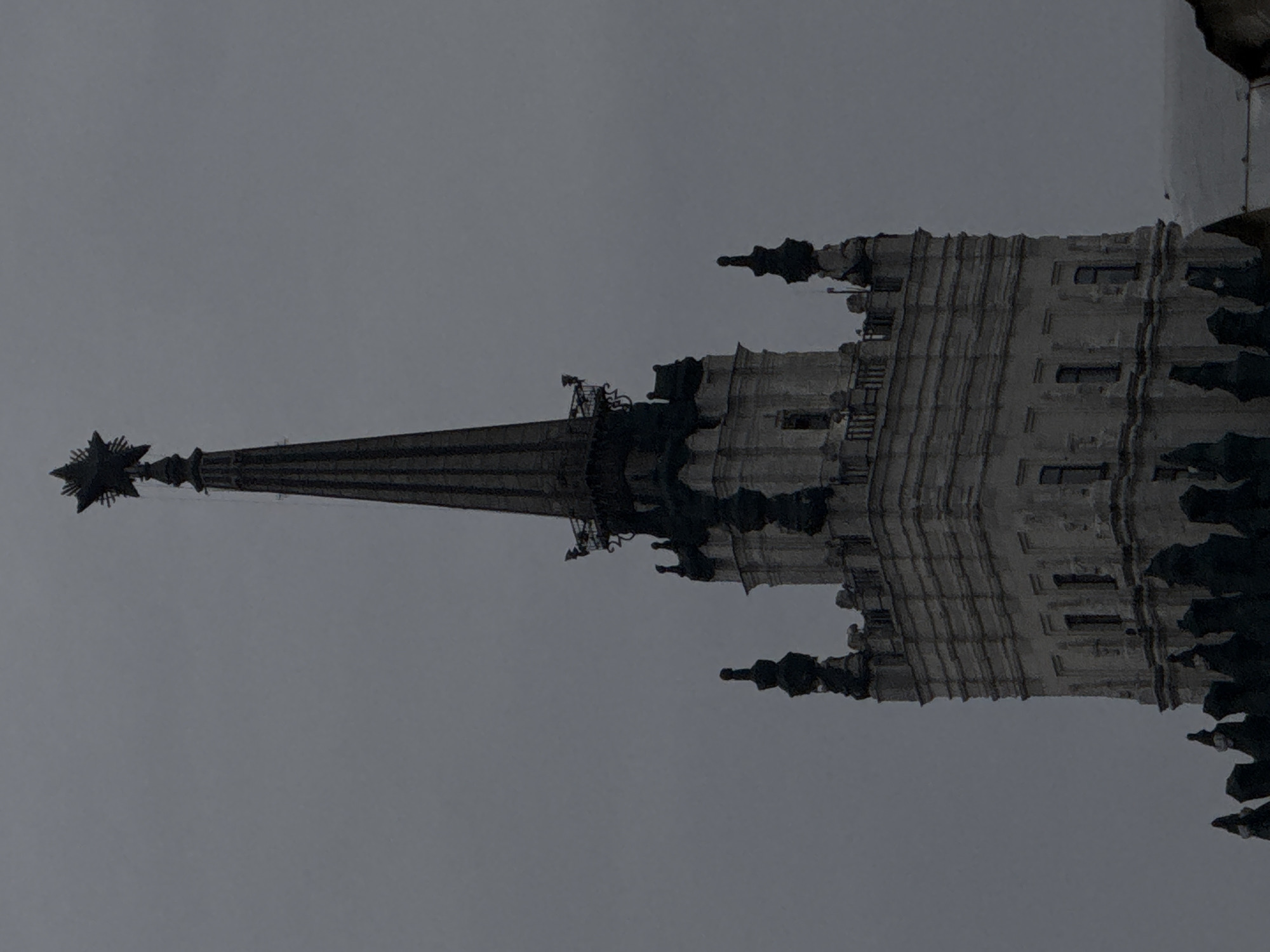 A low-angle shot depicts the upper portion of a Moscow Stalinist skyscraper against a grey sky. The focus is on the central spire, which is topped with a five-pointed star, an emblem from the Soviet era. The architecture features vertical ridges, ornate edges, and small windows. The tower's gothic-inspired design is accented by shadowy gargoyle-like projections along the edges. The color palette is predominantly grey, creating a solemn atmosphere.
