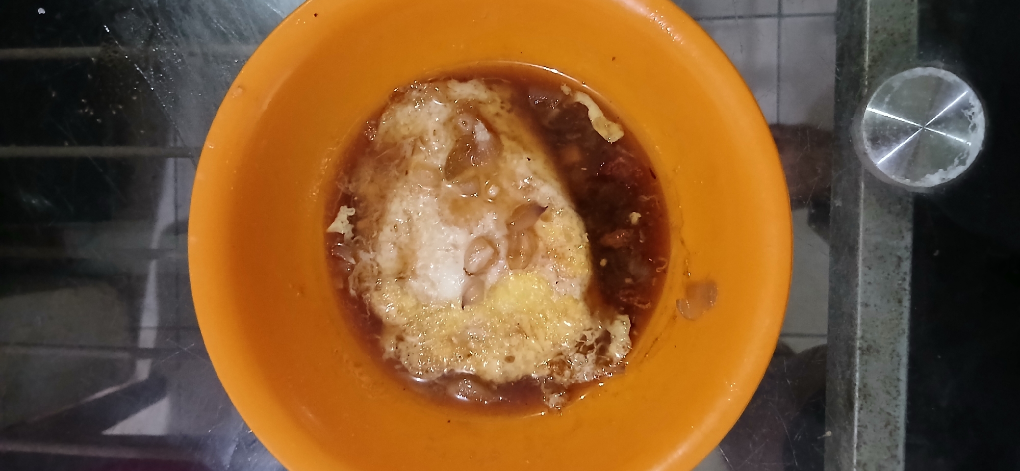In Samarinda, Indonesia, a close-up shot captures an orange bowl filled with food. The bowl contains a fried egg swimming in a dark, savory broth. Scattered around the egg are small pieces of what appear to be garlic or shallots. The bowl rests on a reflective surface, possibly a glass table, which shows streaks and some smudges. The background is blurred but suggests a domestic or casual setting with vertical elements that could be part of furniture or shelving. The lighting is dim, contributing to the overall moody atmosphere of the image.