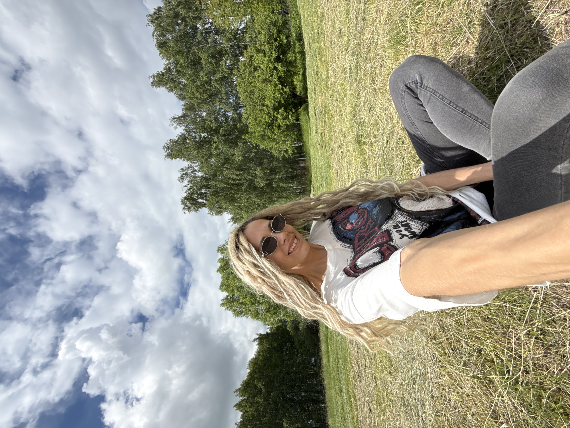 A woman is captured in a selfie outdoors, smiling broadly at the camera. She has long, blonde hair styled in dreadlocks or braids and wears round, dark sunglasses. Her attire consists of a white t-shirt with a colorful graphic design featuring abstract elements and what appears to be upside-down text that might read "You Want", along with dark grey jeans.
The woman is positioned on dry, light brown grass, suggesting a park or an open field. The background features a dense line of tall, green deciduous trees, possibly birch, stretching across the landscape. Above, the sky is bright blue with numerous white, fluffy cumulus clouds.
The image appears to be taken on a clear, sunny day, likely mid-day or early afternoon, given the bright illumination and visible shadows. The woman's expression conveys happiness and relaxation as she enjoys the outdoor setting. The location context for this scene is Rusanovo, Russia.