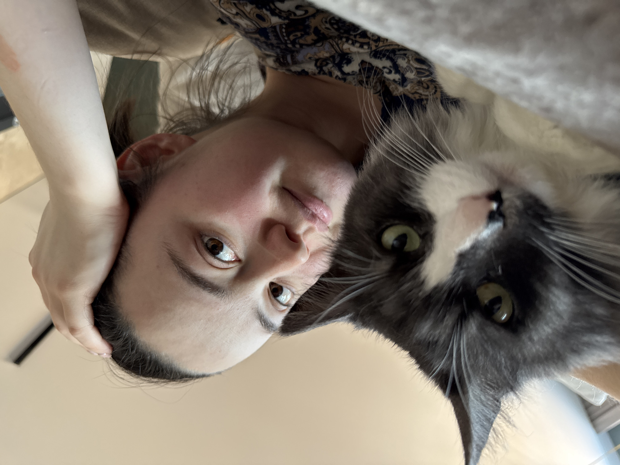 A woman and a gray and white cat are photographed from a low angle. The woman's face is partially visible, with brown eyes and dark hair pulled back. Her right arm is raised, with her hand near her forehead. She is wearing a patterned shirt. The cat's face is prominent, with green eyes and long whiskers. The background is a light beige color, possibly a wall or ceiling. The location is not identifiable as Izhevsk, Russia based on the image content.