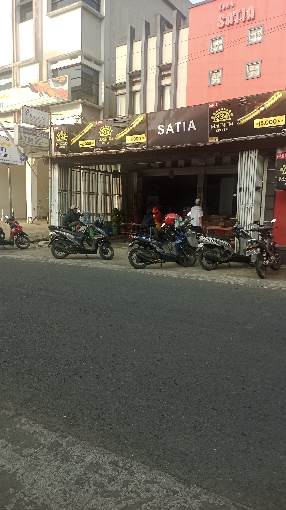 The image shows a street scene in Karyamandala, Indonesia, with a row of shops on the right side of the frame. A multi-story building with a red facade and the prominent sign "SATIA" in large pink lettering is visible at the top right. Below this building, a storefront with an open entrance is flanked by metal shutters. Several motorcycles are parked along the sidewalk in front of the shops.
A banner advertising "234 MAGNUM KRETEK" with prices is displayed across the storefront. Inside the shop, people are visible, including one wearing a white shirt and cap, and another in a red shirt and yellow helmet. Gas tanks are stacked near the entrance. A person wearing a white cap is sitting on one of the motorcycles, holding a large water bottle.
The street in the foreground is paved with asphalt. The overall lighting suggests it is daytime, with sunlight casting shadows. Signs for "SALON" and "ATM" are visible on a building to the left, indicating a commercial area.