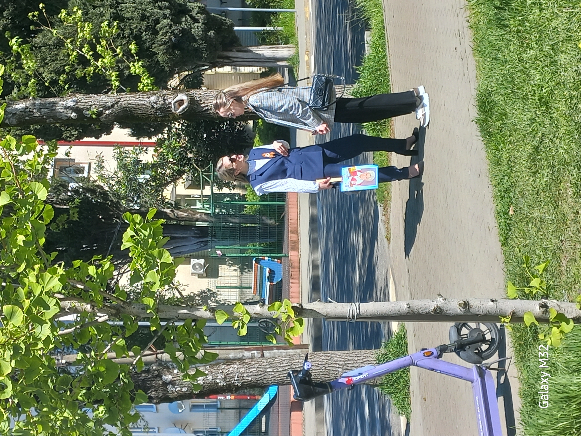 Here is a description of the image:
Two women are walking on a sidewalk in Sochi, Russia. The woman closer to the camera is wearing a blue suit and holding a small portrait. She also has an orange ribbon pinned to her lapel. The other woman has long hair and is wearing a gray jacket, black pants, and white sneakers. There is a scooter parked near a tree on the sidewalk. The background includes trees, grass, and buildings.