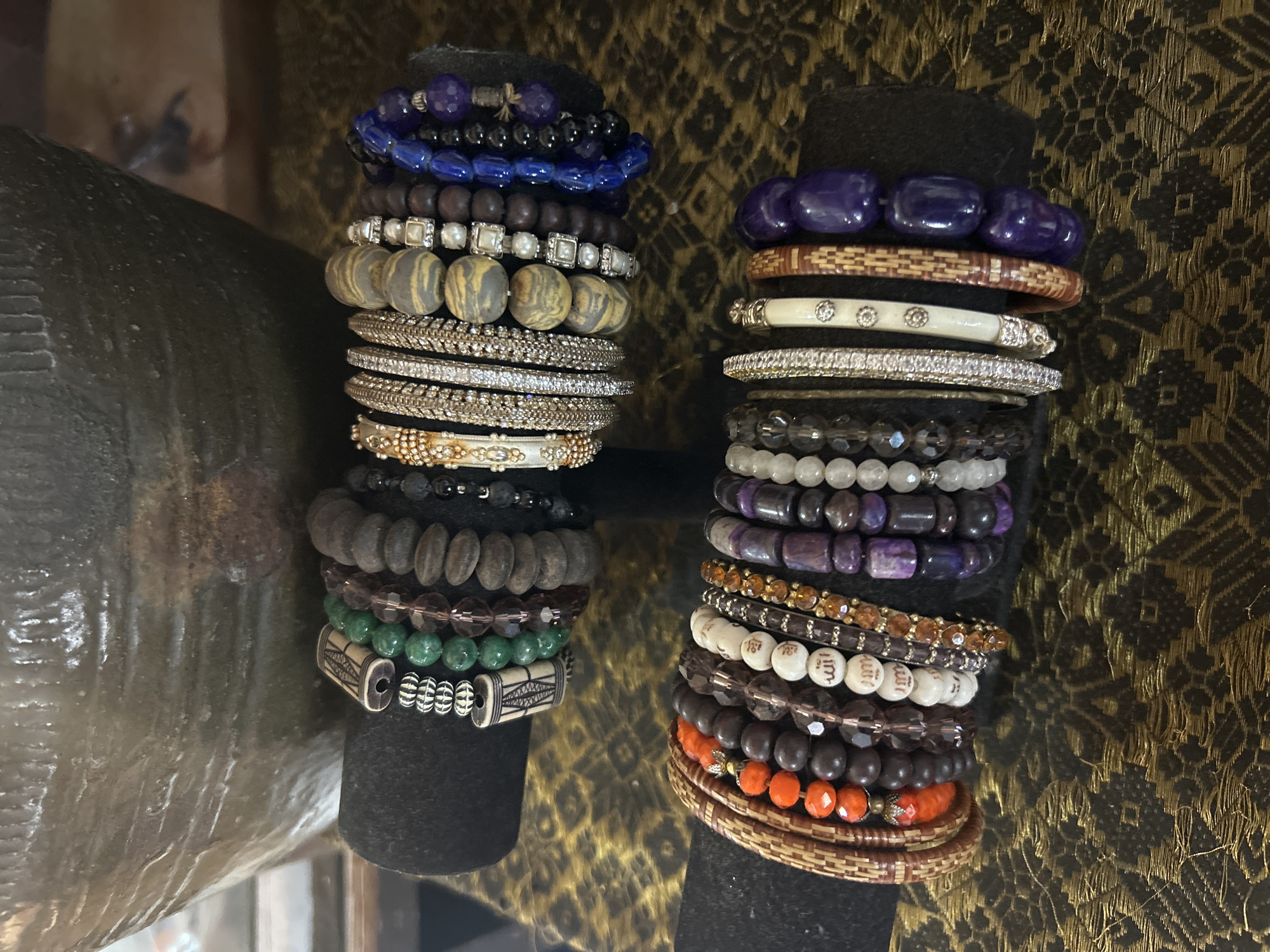 A close-up, eye-level shot features two black velvet bracelet stands displaying an assortment of bracelets. The stands are placed on a patterned gold and black textured surface. Behind the stands, a large, textured, dark brown vase is partially visible. The bracelets are diverse in material, color, and style, ranging from beaded bracelets with gemstones and wooden beads to metal bangles and bracelets with engraved charms.
The top stand showcases bracelets with green jade beads, brown wooden beads, and silver-toned metal accents. Notable among these is a bracelet with a carved bone bead. To the right, several bracelets with a striped pattern of grey and yellow beads are displayed, followed by dark blue and purple gemstone bracelets.
The bottom stand displays bracelets made of interwoven brown material, orange beads, dark brown wood beads, and clear faceted beads. Several white beads with Arabic script are visible on one bracelet. This stand also features bracelets with purple and dark blue beads, culminating in a bracelet of large, smooth, deep purple beads on the right. The lighting is soft and ambient, highlighting the textures and details of the bracelets. The image does not provide any information to indicate its location in Denpasar, Indonesia.