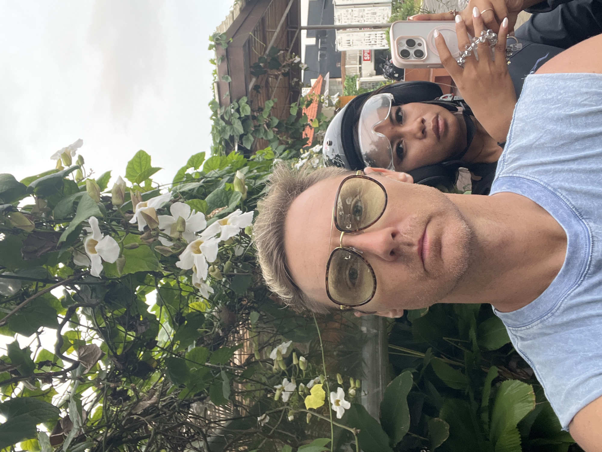 The image shows two individuals taking a selfie. The person in the foreground is a fair-skinned male wearing sunglasses and a blue t-shirt. Behind him is a darker-skinned individual wearing a motorcycle helmet with a clear visor, holding a smartphone with multiple lenses visible on the back. They appear to be riding on a vehicle, possibly a scooter or motorcycle.
The background shows dense green foliage with white flowers. In the blurred distance, there are buildings and structures that suggest an urban or semi-urban environment. The location is identified as Ungasan, Indonesia based on geotags.