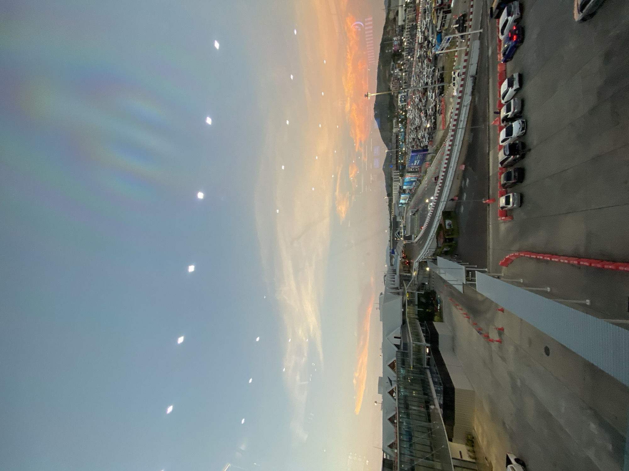 The image shows an aerial view in the city of Mai Khao, Thailand. A sprawling parking lot with numerous cars fills the background, extending toward a mountain range under a partly cloudy, orange-tinted sky at dusk. In the foreground is a racetrack, marked by red cones and a barrier. A row of parked cars lines the edge of the track closest to the viewpoint. Buildings with reflective windows are visible near the track. The image appears to have been taken from an elevated location, potentially a high floor of a building.