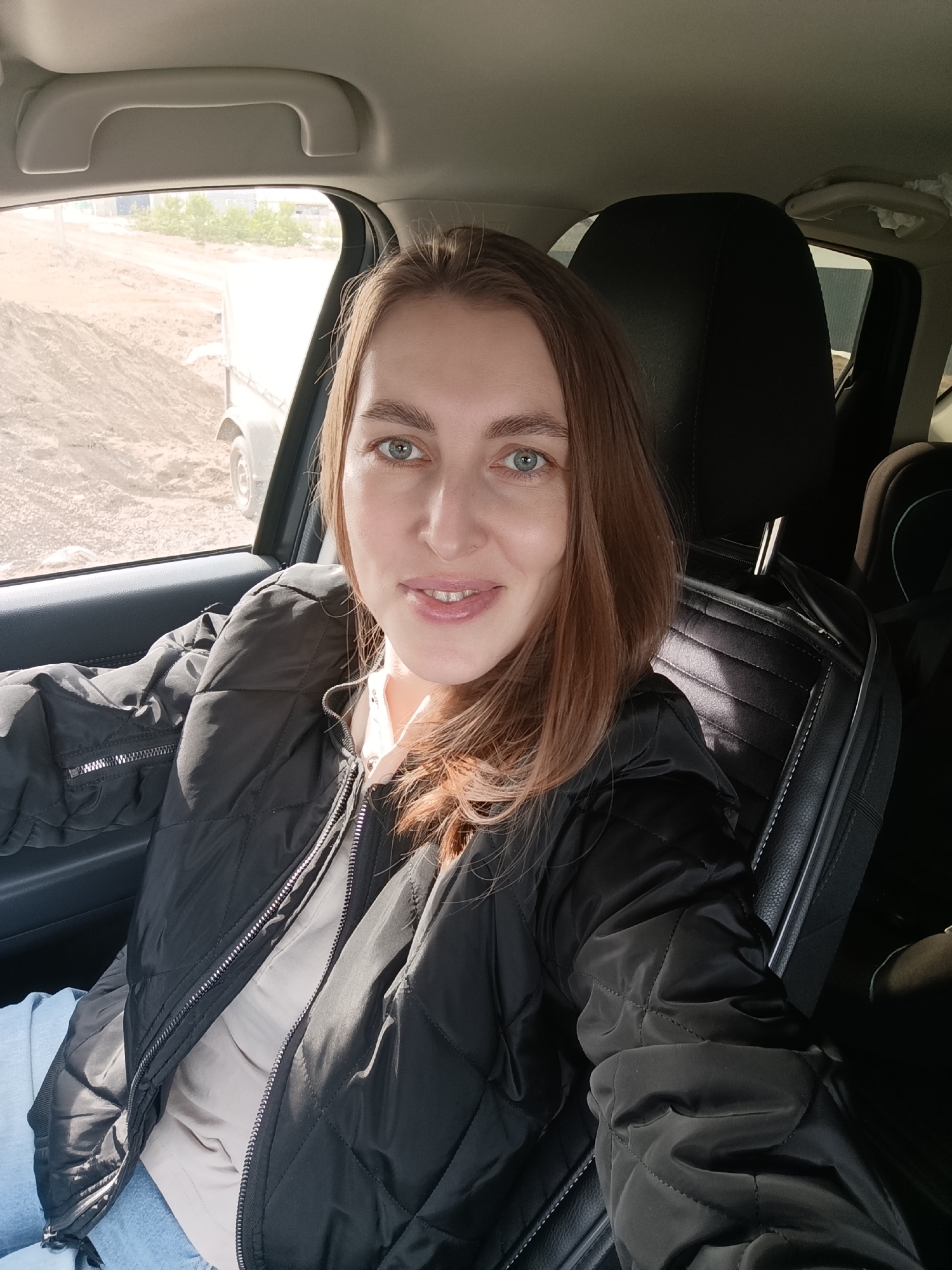 A woman is sitting in the driver's seat of a car, smiling at the camera. She is wearing a black quilted jacket over a light-colored shirt. The interior of the car is visible, including the seat headrest and the car door. Outside the car window, a construction site with a white truck and piles of dirt is visible. Without additional data such as GPS metadata embedded in the image or associated geotagged social media posts, providing a forensic level location detail to Chaikovsky, Russia is impossible.