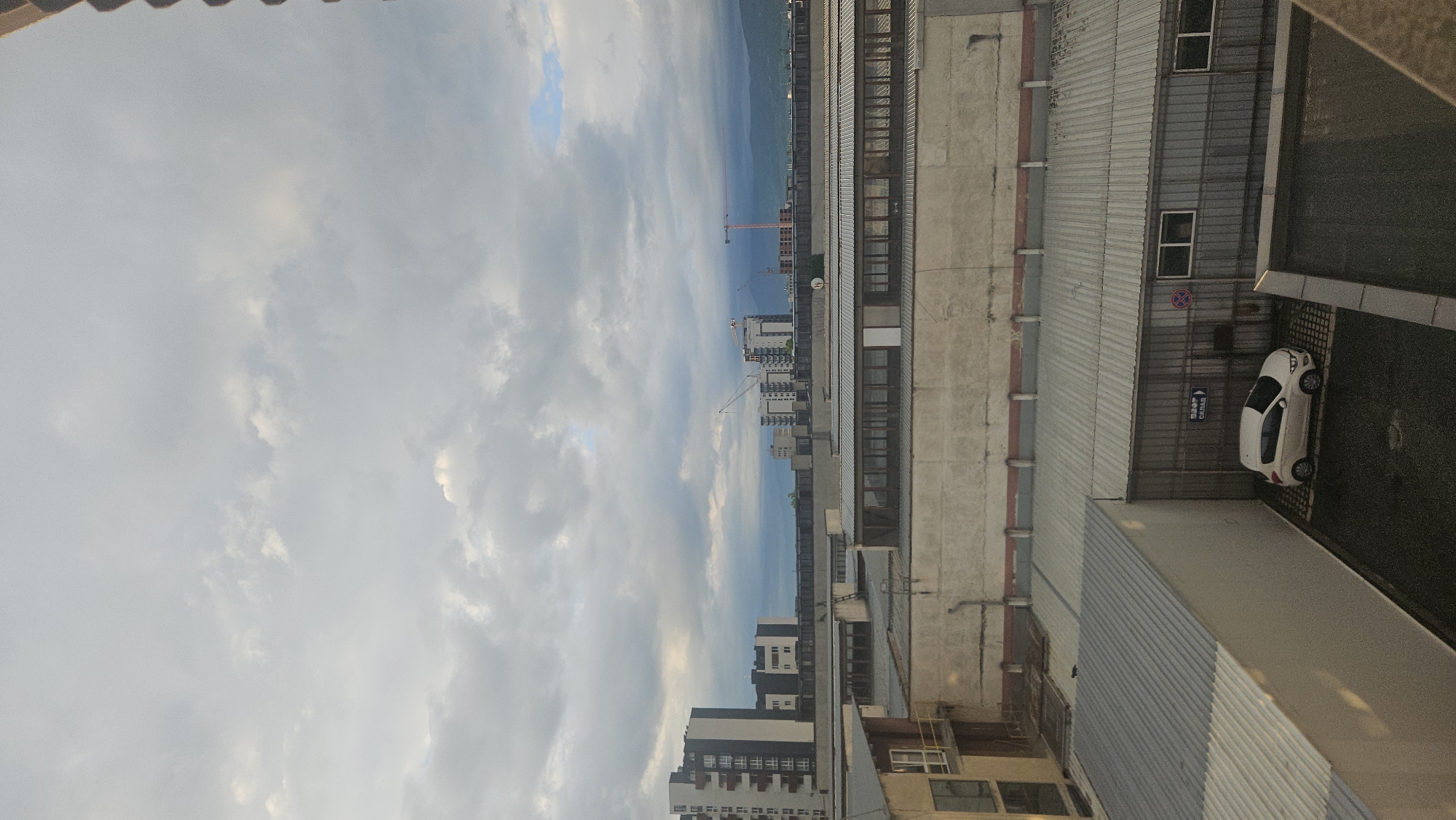 The image is an elevated view of Plovdiv, Bulgaria, with the sky and a cloudscape dominating the left side of the frame. In the center are some buildings.
In the bottom right corner is a white car parked in a lot next to a row of industrial buildings. A sign with text in Cyrillic is mounted on the wall near the car.
To the right of the cityscape are mountain ranges.
The image appears to have been taken from a high-rise building or apartment window.