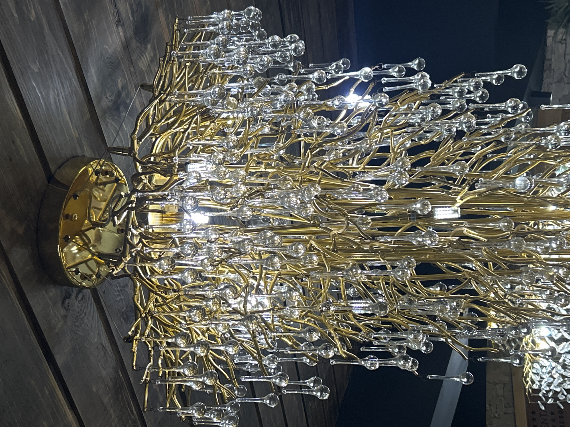 This image shows a close-up, low-angle shot of a large, ornate chandelier. The chandelier is gold-toned and features a complex arrangement of thin, branching structures from which numerous clear, teardrop-shaped glass ornaments are suspended. The ceiling above is made of dark wooden planks. The background is mostly dark, suggesting an indoor setting at night. No people are visible in the image. The chandelier is the sole subject of the photograph.