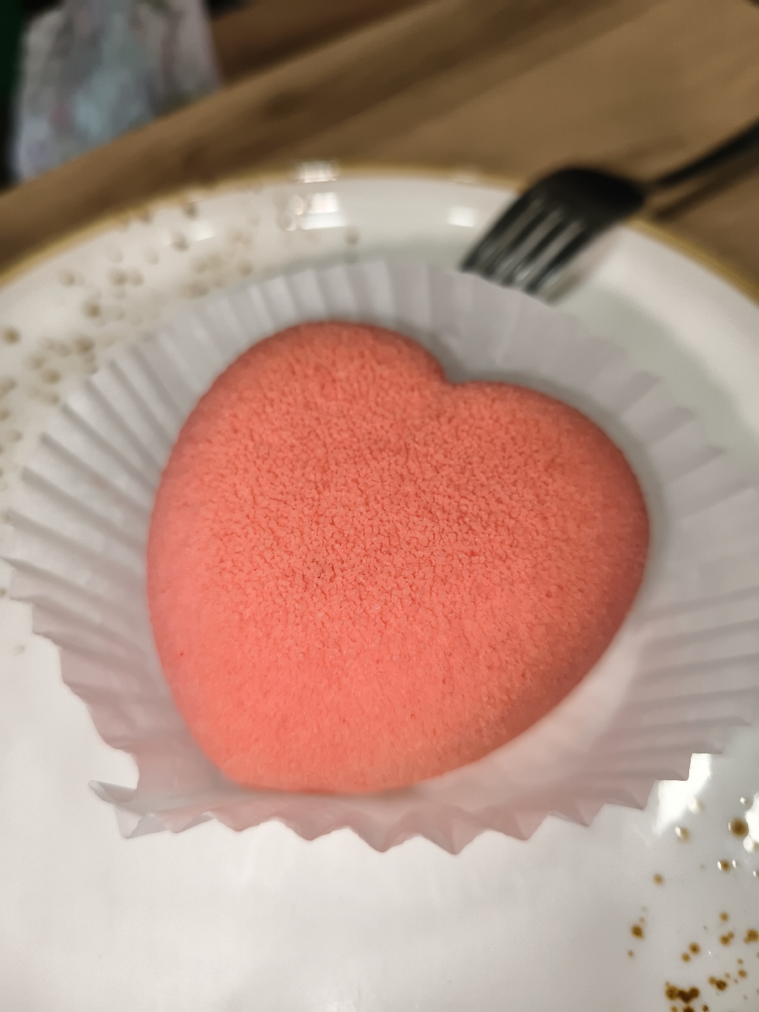 A heart-shaped, pink, mousse-like dessert sits on a white paper liner, which is placed on a white plate. The dessert has a fine, granular texture. A black fork is visible in the upper right corner of the frame, positioned above the plate. The background is out of focus, showing a wooden surface and possibly some other objects. The scene appears to be indoors, likely a cafe or restaurant. There are no people visible in the image.