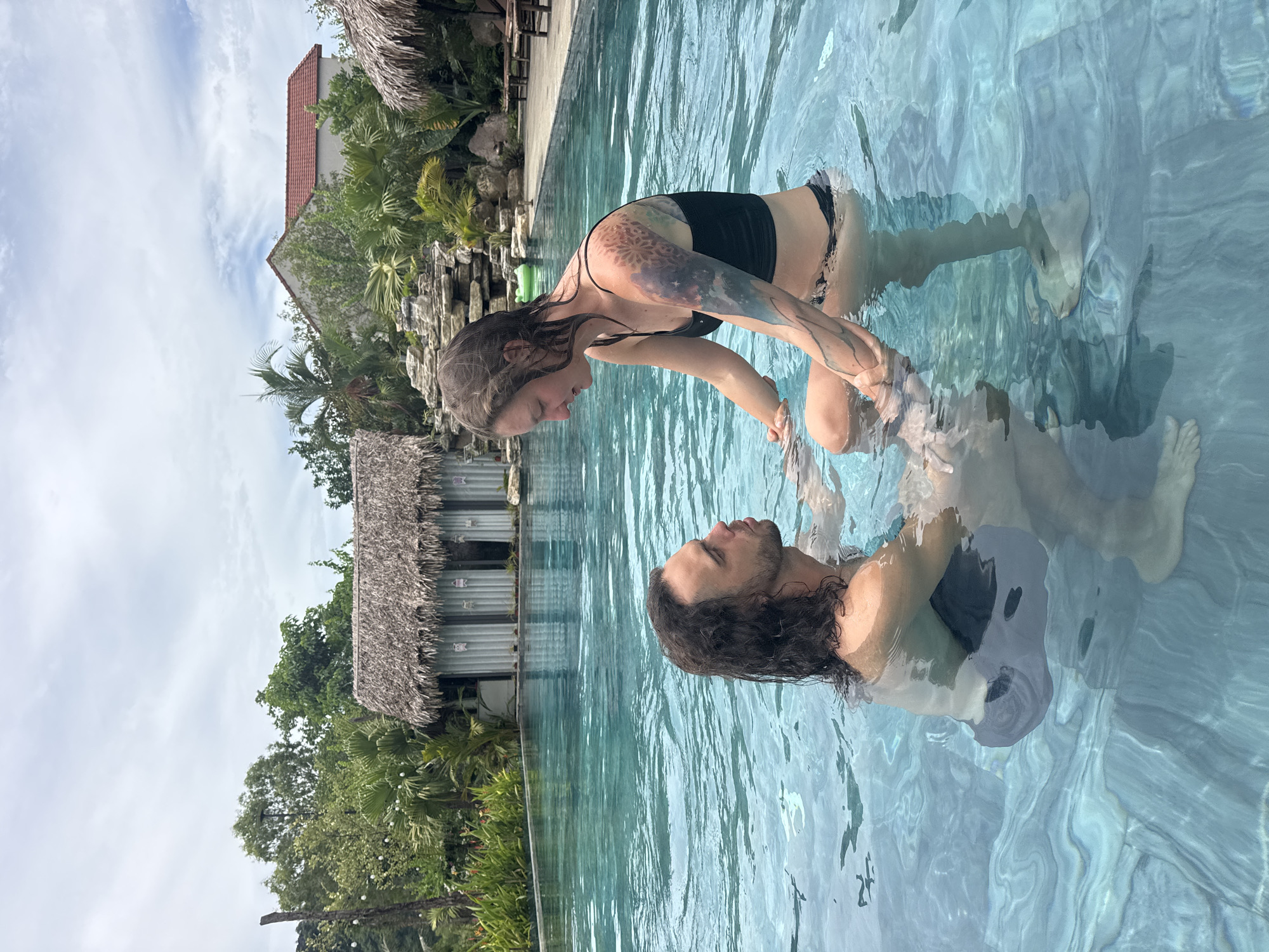 An outdoor infinity pool in Nam Hoa Lư Ward, Vietnam, features a couple submerged in the turquoise water. The man, with long dark hair, faces the woman, who is seated with her legs bent. She is wearing a black bikini top and bottoms, and has a large tattoo on her left arm. They are holding hands and looking at each other. In the background, there are several thatched-roof buildings, lush green trees, and a cloudy sky. The pool's floor is visible through the clear water, showing a patterned surface.