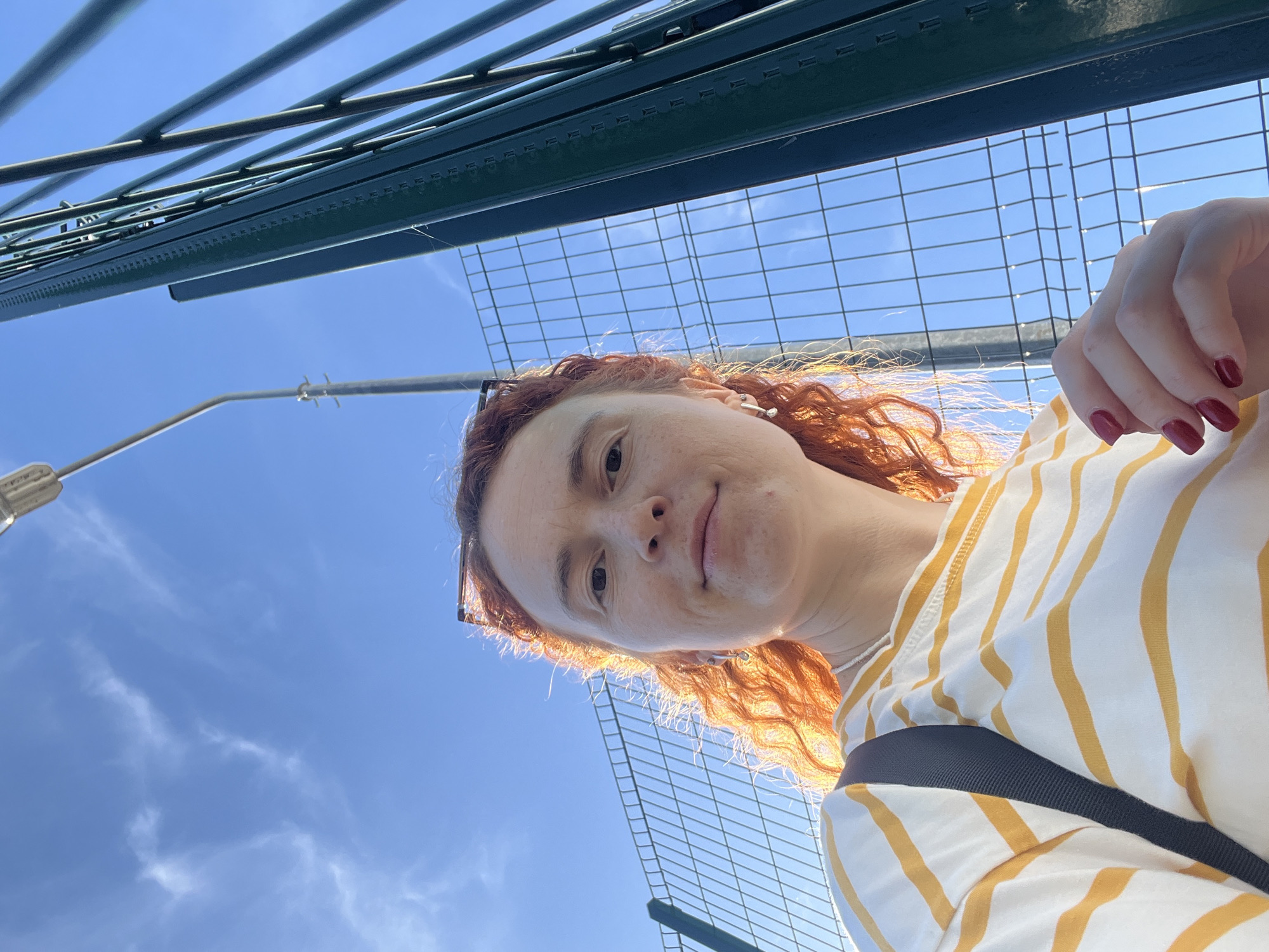 A selfie taken outdoors, looking up at a woman with red curly hair. She is wearing a white and yellow striped shirt with a black strap across her shoulder. Her fingernails are painted dark red. Above her are a chain-link fence, a green metal structure, and a street light pole. The sky is blue with some wispy clouds. It is daytime. The location is in the city of Chaikovsky, Russia.