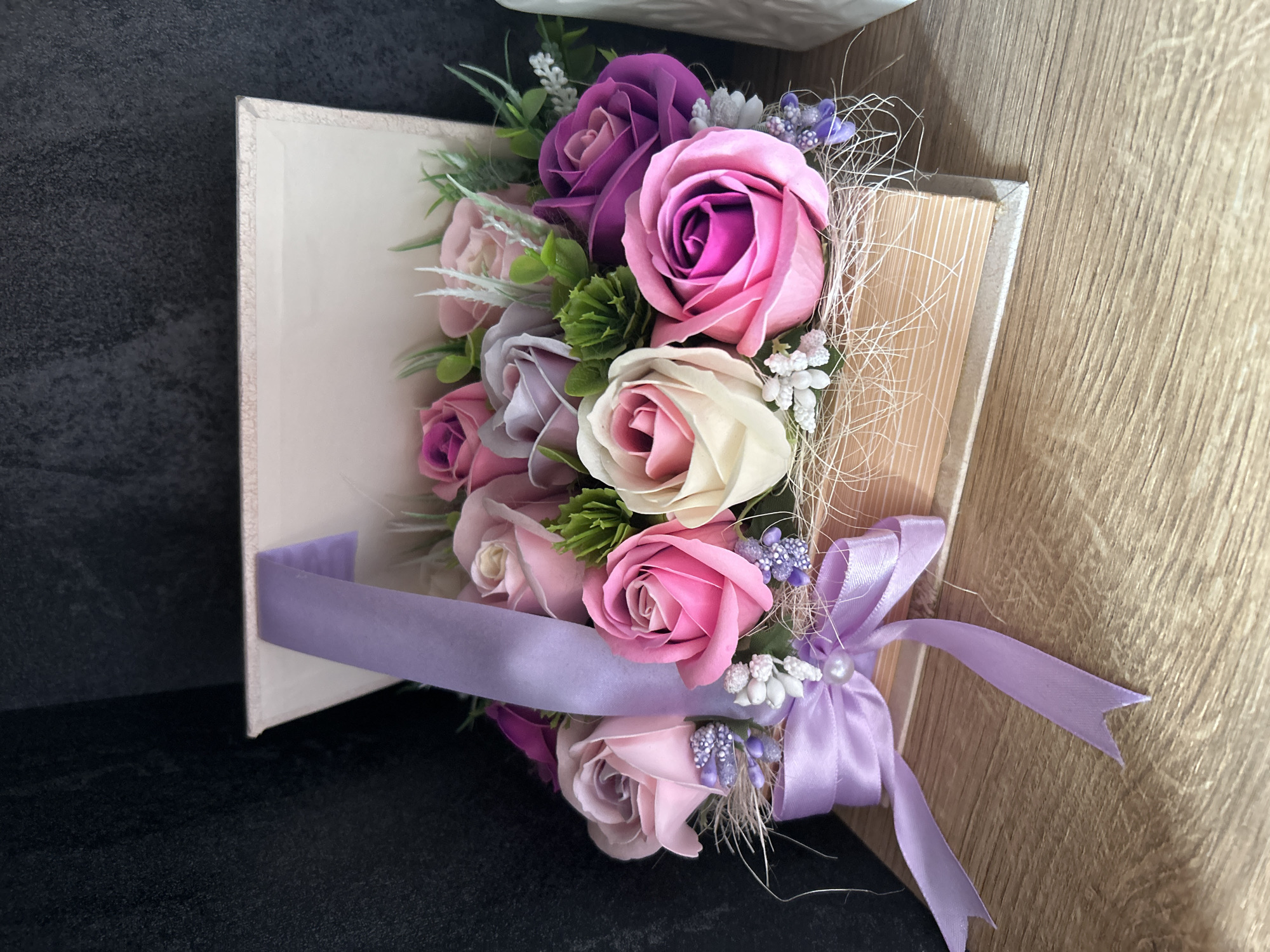 This eye-level, close-up shot features a beautifully crafted decorative arrangement of artificial flowers, primarily roses, presented as if bursting from a light-colored book-shaped box. The "book" stands upright, showcasing its spine on the left and its textured "pages" side on the right.
The floral arrangement is a vibrant mix of artificial roses in various shades of pink, ranging from deep fuchsia and bright pink to softer pastel hues, complemented by lavender, light purple, and creamy white blossoms. These roses are accented with artificial green foliage and small clusters of tiny white and lavender berry-like elements, adding depth and texture. Fine, shredded, light-colored material, possibly raffia, is woven throughout the bouquet, further enhancing its visual appeal.
A wide, satin ribbon in a soft lilac shade is elegantly wrapped around the lower portion of the book and tied into a graceful bow on the right side. A small, lustrous pearl-like bead is fixed at the center of the bow, adding a delicate touch.
The decorative piece rests on a dark, textured surface on the left, likely a table or shelf. To the right, a warm, light brown wooden surface provides a complementary background. The lighting suggests an indoor setting, highlighting the intricate details and harmonious color palette of the arrangement. There is no visible text within the image.