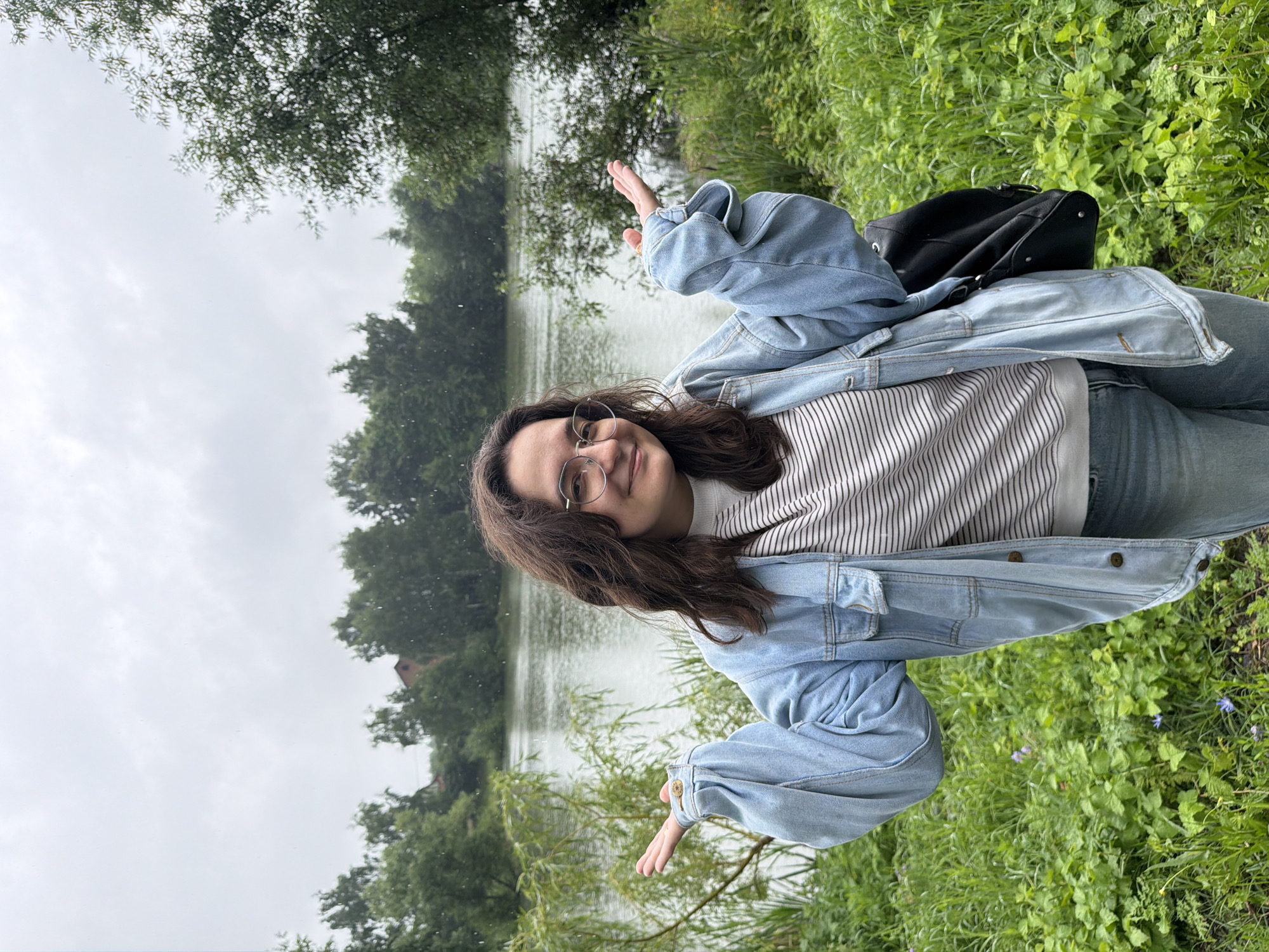 A young woman with dark, wavy hair and glasses stands in front of a body of water, possibly a lake or river, in Nikulskoye, Russia. She is wearing a light blue denim jacket over a striped long-sleeved shirt and blue jeans. Her arms are outstretched, palms up, in a gesture that could be interpreted as questioning or welcoming. A black backpack is slung over her right shoulder. The background consists of lush green vegetation and trees, with a slightly overcast sky above. The overall impression is that of a casual outdoor setting.