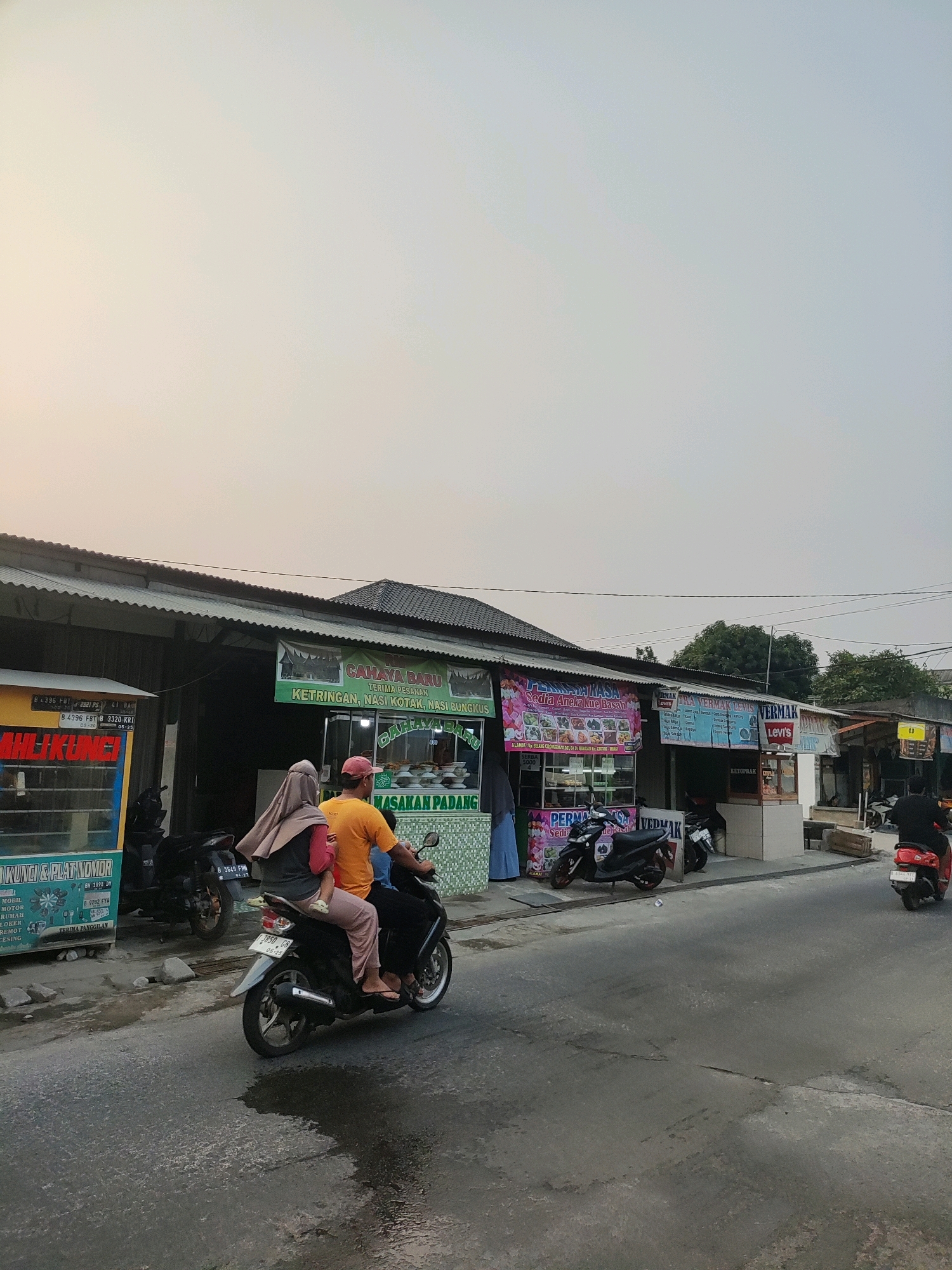 The image depicts a street scene in Wanajaya, Indonesia, at a location with several small businesses. In the foreground, a black motorcycle is traveling away from the viewer. On the motorcycle are three people: an adult male driver wearing an orange t-shirt and a red cap, an adult female passenger wearing a pink headscarf and a grey shirt, and a child seated in front of the female passenger. The motorcycle is on a paved road with some wet patches.
To the left of the road is a row of single-story buildings with corrugated metal roofs. Several of these buildings house small businesses. One business has a yellow and blue facade with text that appears to be "AHILIKUNCI" and "PLAT NOMOR". Another business has green and white patterned walls and a sign that reads "MASAKAN PADANG" and "CAHAYA BARU" with text indicating "KETRINGAN, NASI KOTAK, NASI BUNGKUS". Next to this, another business displays a pink banner advertising "PERMA KASA SEDIA KUE BESAR". Further down the row, a business with a white facade has signage for "VERMAK LEVIS".
Several other motorcycles are parked in front of the businesses. In the background, more buildings and trees are visible. The sky is overcast with a soft, hazy light. The overall impression is of a modest commercial area in a tropical or subtropical setting.