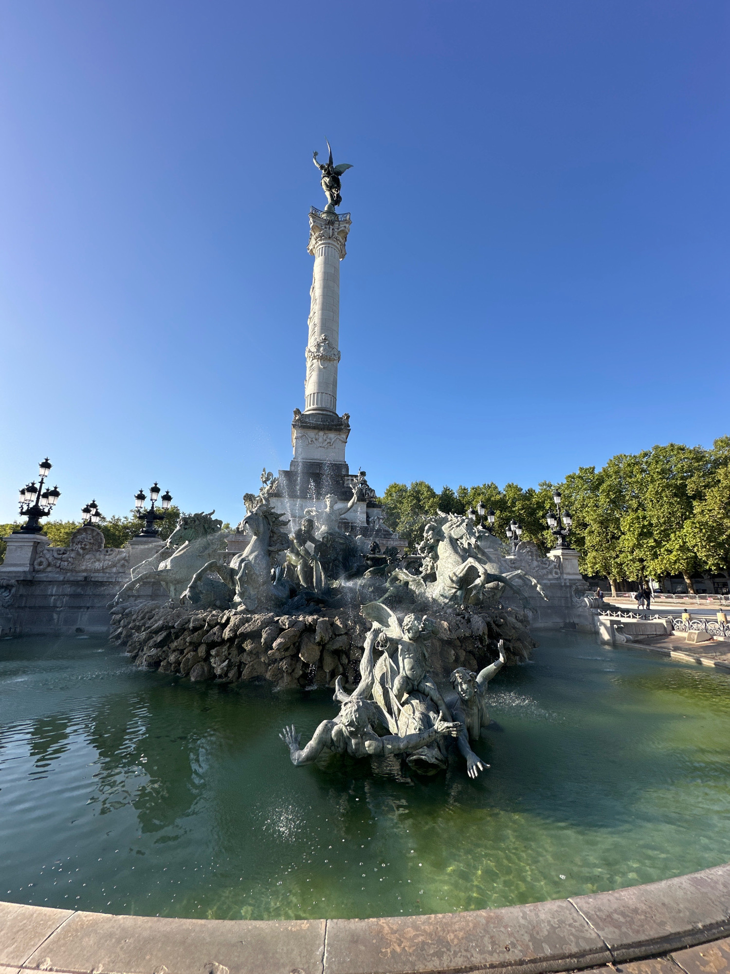 The image shows the Place de la Victoire in Bordeaux, France, featuring the Monument aux Girondins, a grand column topped with a statue of Liberty breaking its chains. The monument is surrounded by an elaborate fountain with bronze sculptures of mythological figures, horses, and other creatures, with water cascading and spraying. In the foreground, the fountain pool is a deep green, reflecting the sky and the surrounding greenery. Beyond the fountain, lush green trees line the square, and ornate lampposts are visible. In the background, several people are seen walking on a paved area. The sky is clear and bright blue.