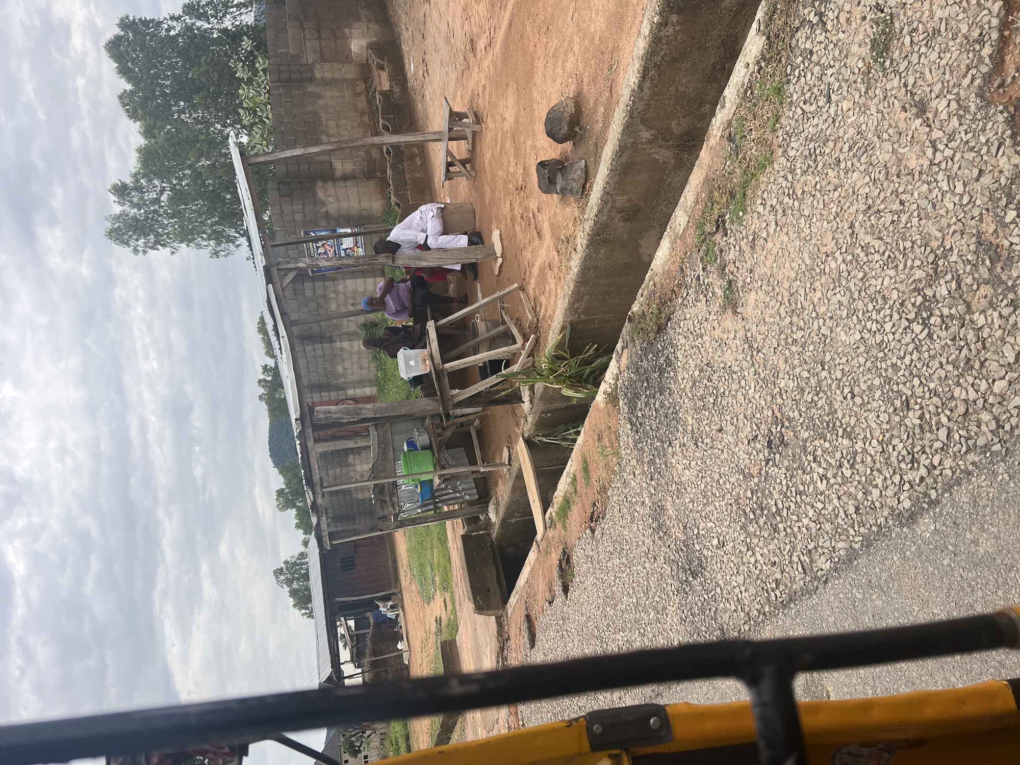 The image depicts a scene in Jalingo, Nigeria, possibly on an overcast day. In the foreground, a yellow structure, likely part of a vehicle, is visible. The main subject is a rustic wooden structure with a corrugated metal roof, serving as a shelter. Beneath this shelter, several people are gathered, appearing to be engaged in conversation or taking a break. One person, dressed in white, is seated on a wooden block, while others are seated on a wooden bench or the ground.
The area around the shelter is dusty and uneven, with some patches of green grass. To the right of the shelter, a concrete drainage channel runs alongside a gravelly path. Behind the shelter, a brick wall and more wooden structures are visible. Trees with lush green foliage are seen in the background, and a hill can be faintly observed in the distance.
The overall atmosphere seems relaxed and informal, suggesting a casual gathering or a stop along a route. There are no explicit indications of a specific event, but the presence of people under a shelter suggests a pause in their day. A poster with some text and images is affixed to the wall behind the people, but the text is illegible. The lighting suggests it might be late morning or afternoon, given the overcast sky.