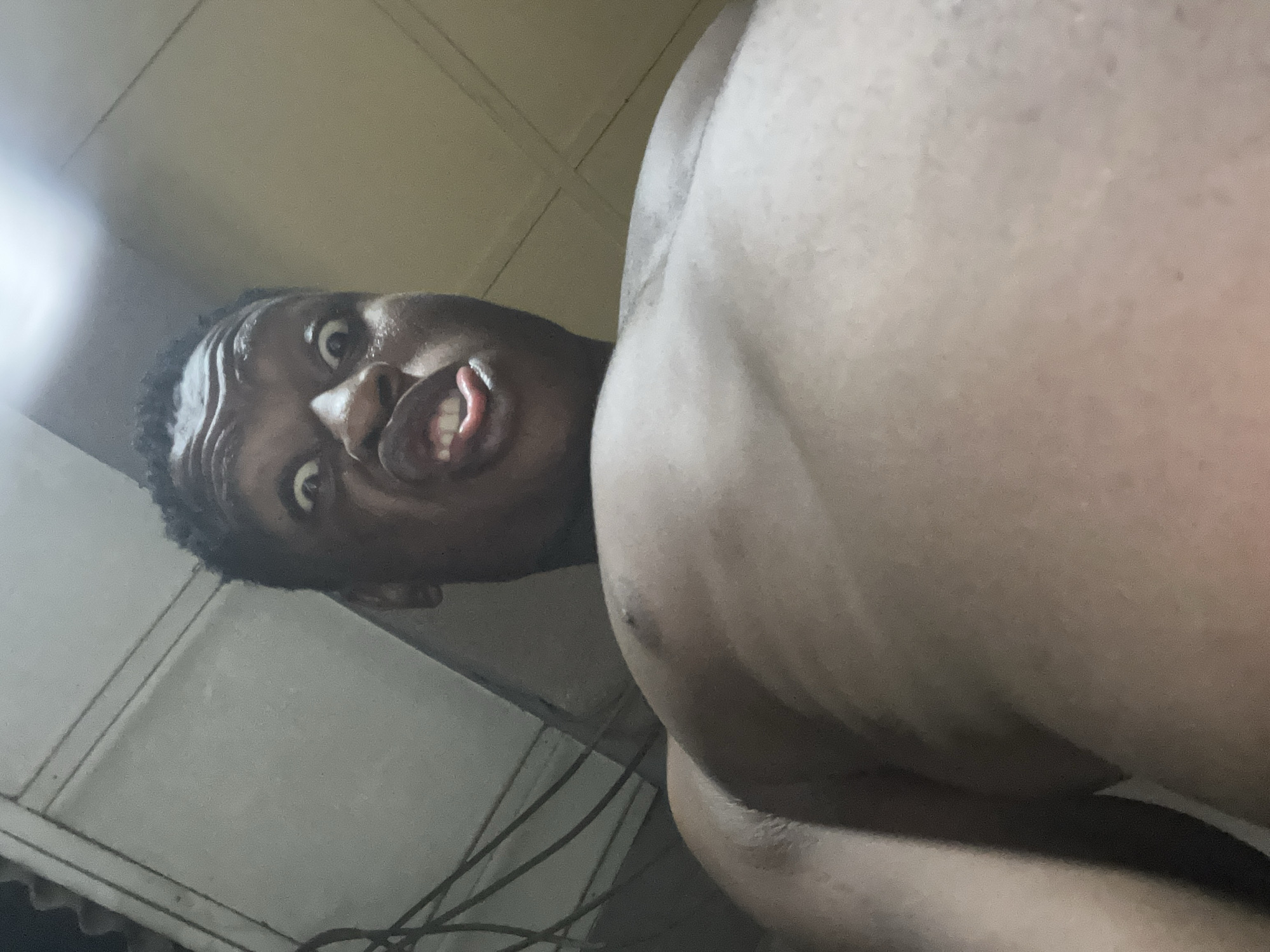 An adult male with dark skin and short, dark hair is depicted from a low-angle perspective, looking upwards at the subject. The subject is shirtless, revealing their upper chest and shoulders. Their face displays an exaggerated expression with wide-open eyes, furrowed brows, an open mouth, and a partially extended tongue. The background indicates an indoor setting, featuring a light-colored ceiling, possibly paneled, with dark electrical wires visible in the upper left quadrant. A bright artificial light source is positioned overhead in the upper central-right of the frame, illuminating the scene. This scene takes place within an unspecified indoor location in Ikorodu, Nigeria.
