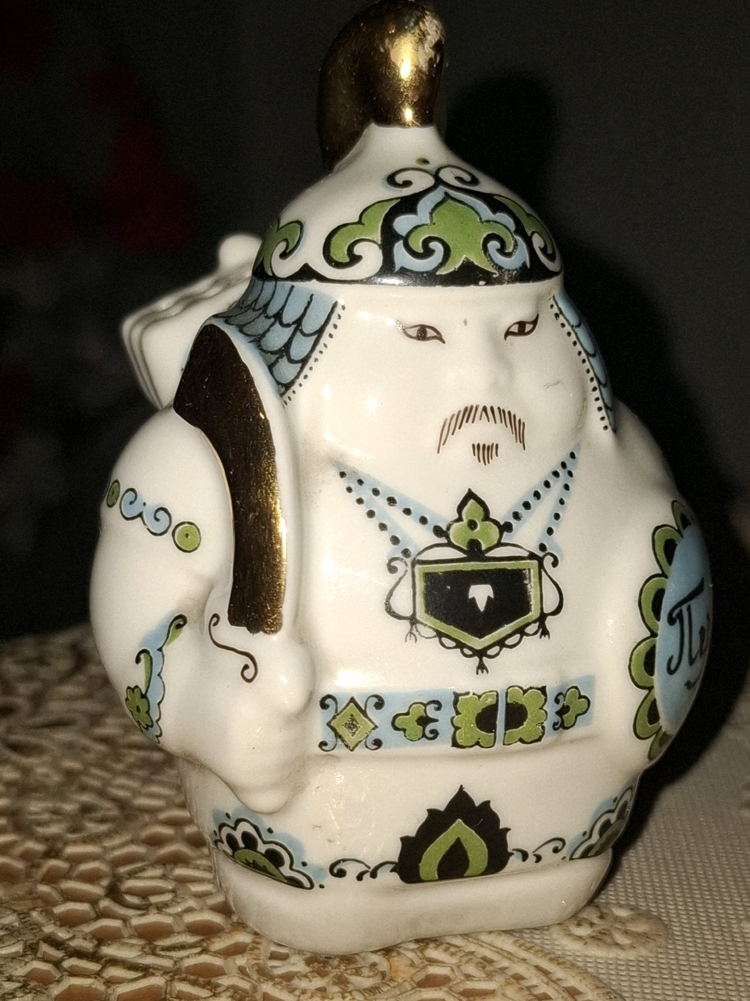 A close-up, high-angle studio photograph shows a ceramic figurine of a stylized warrior or historical figure. The figurine is predominantly white with intricate blue, green, and gold detailing. It has a round face with painted brown eyes and a thin mustache. The figure wears a helmet with a gold finial, ornate patterned clothing, and appears to be carrying a bag over its left shoulder. The background is dark and out of focus, with the figurine positioned on a textured surface, possibly a doily or tablecloth, with a honeycomb-like pattern. The lighting creates reflections on the glossy surface of the ceramic. The image does not contain any discernible elements that would allow for its location to be identified within Almaty, Kazakhstan or any other specific geographic location.