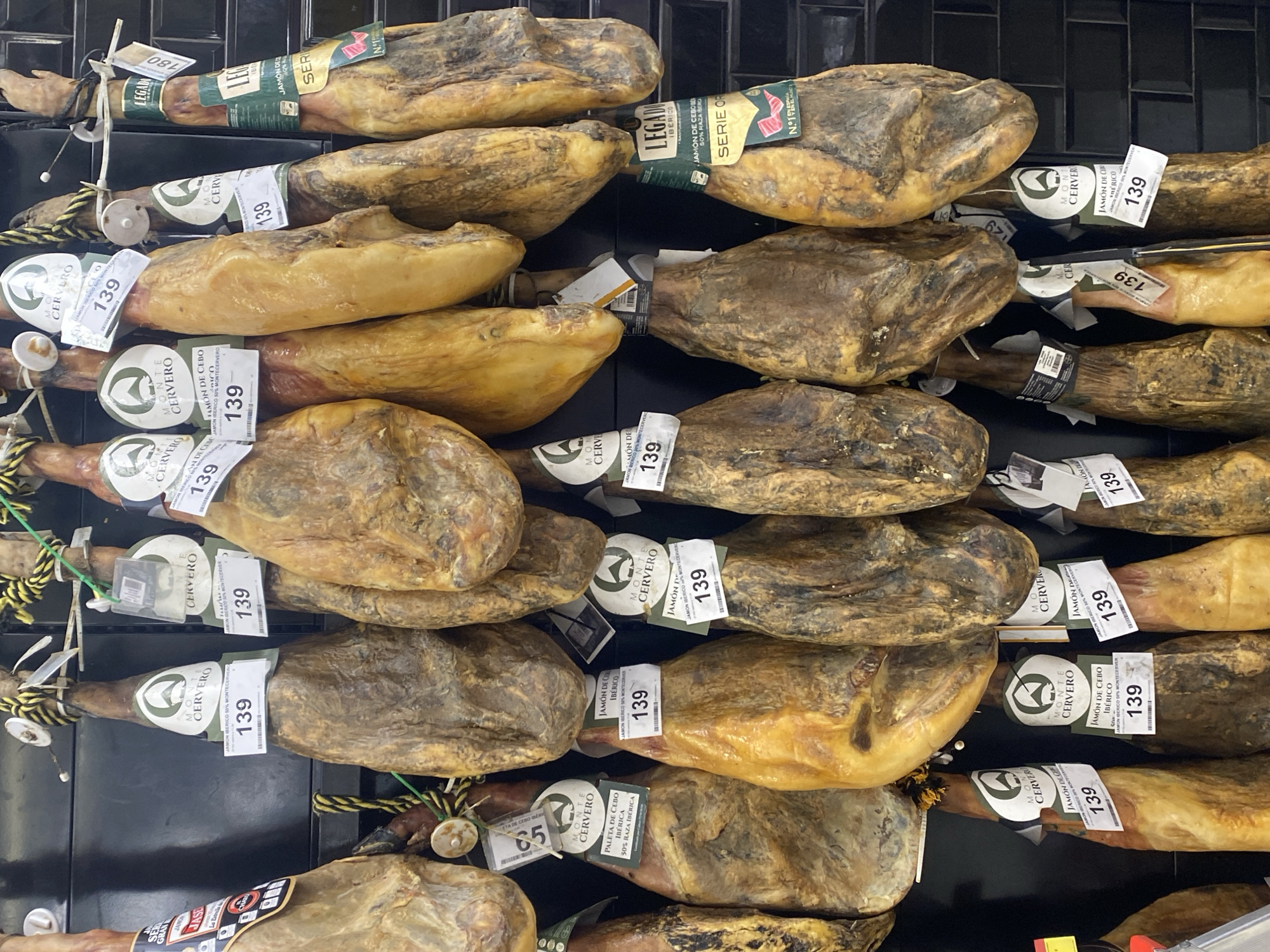 The image depicts a retail display of various cured hams, likely jamón ibérico, hanging in rows. The hams are brown and have a textured, cured appearance. Many of the hams are adorned with labels, some of which display prices, such as "139" and "180," and branding like "MONTE CERVERO" and "LEGADO IBÉRICO." The background consists of a dark tiled wall. The scene suggests a food market or a specialty store in Bel Air, Spain, where these hams are presented for sale. No people are visible in the image.
