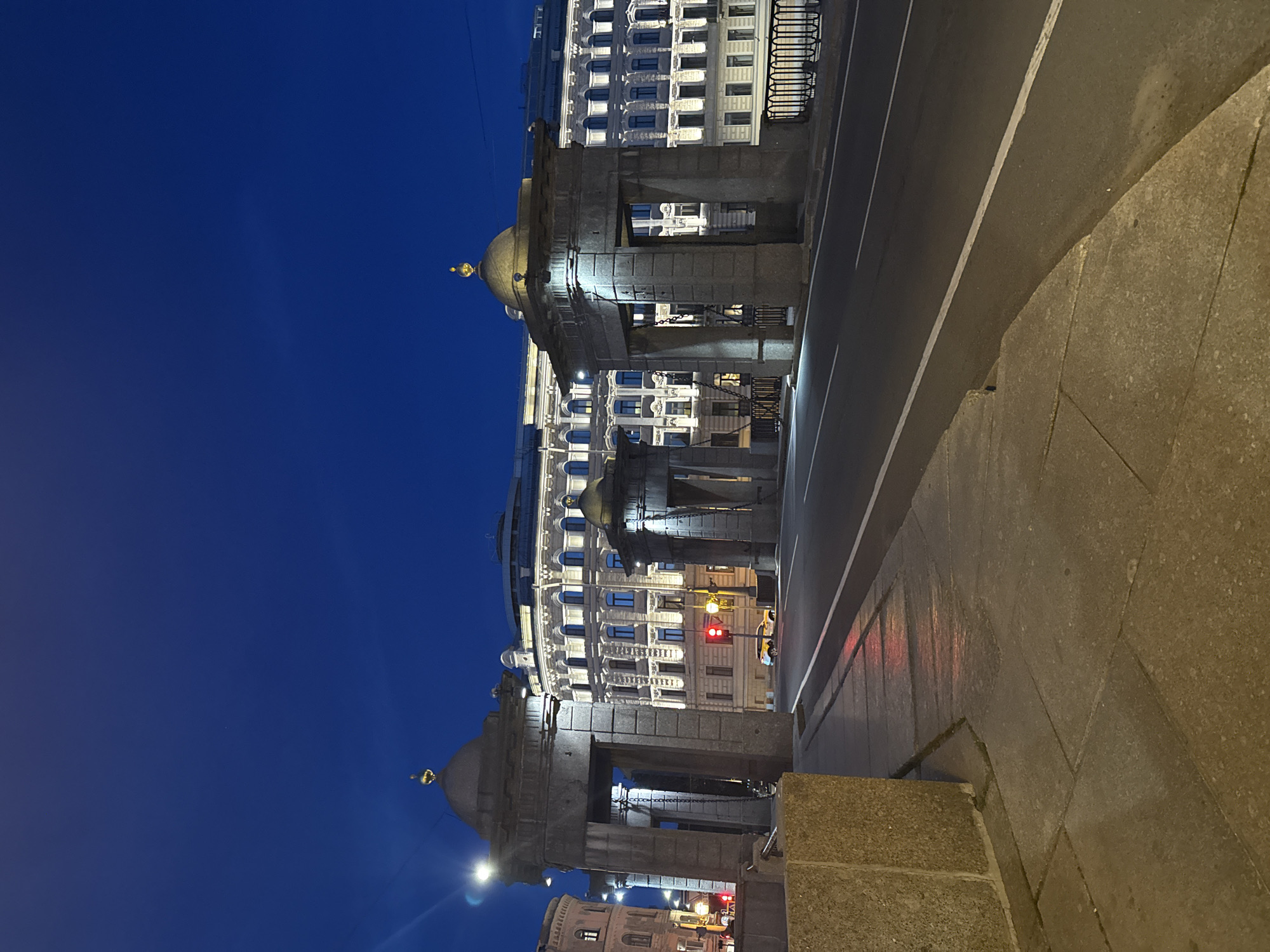 This is a nighttime, eye-level, full shot of the Bank Bridge in Saint Petersburg, Russia. The bridge is illuminated by artificial lights, casting reflections on the wet pavement. The sky above is a deep, clear blue, indicating twilight.
In the foreground, a stone sidewalk is visible, with a concrete barrier on the left. A road runs through the center of the frame, marked with white lines. On the right side of the road, a decorative black metal fence lines the bridge.
The middle ground features the distinctive granite archways of the Bank Bridge. These archways are topped with domes that have small golden spires. Chains hang from the archways, a characteristic feature of the bridge's design.
In the background, a grand, light-colored building with numerous windows is brightly lit, highlighting its architectural details. A yellow car is visible on the road, and a red traffic light is illuminated. The overall scene conveys a sense of the historic and illuminated cityscape of Saint Petersburg at night.