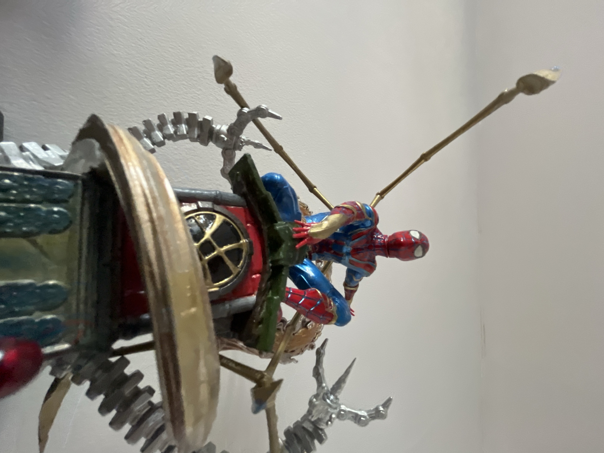 The image shows a close-up, low-angle shot of a Spider-Man action figure on a multi-tiered display platform. The figure is red and blue with gold accents, posed in a sitting position. The platform is elaborate, featuring metallic mechanical arms, gears, and decorative elements in red, gold, and green. The background is a plain, light-colored wall. The scene is indoors, with no visible context of Shumen, Bulgaria.