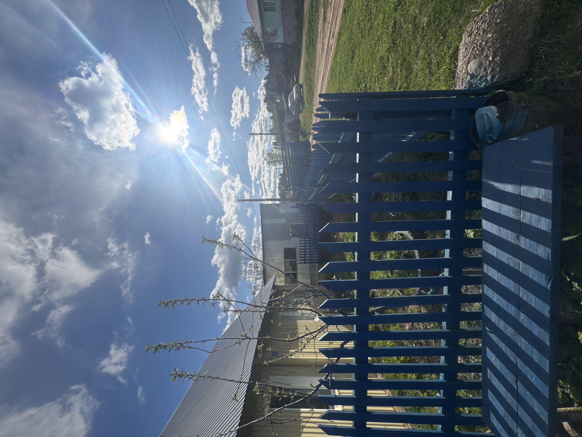 The image captures a bright, sunny day in Ol'khovochka, Russia. The scene is dominated by a vivid blue wooden fence in the foreground, casting strong shadows. Beyond the fence, a residential street scene unfolds. Houses are visible, with one painted yellow with a corrugated metal roof partially obscured by a budding tree. A dirt road runs along the edge of the property, with greenery lining the side. A car is parked further down the road, in front of another house with a green exterior. In the top right corner, a large, gray stone is situated near the fence with an old barrel and a blue plastic container beside it. The sky is a brilliant blue dotted with fluffy white clouds, and the sun shines intensely, creating lens flare.