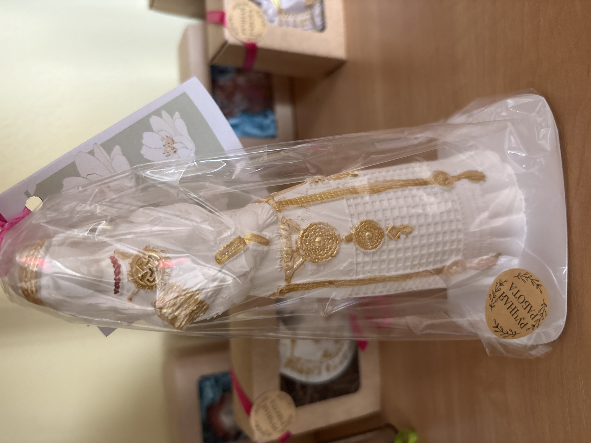 A close-up, eye-level shot shows a white, molded candle in the shape of a figure adorned with gold accents. The candle is encased in clear plastic packaging, tied at the top with a pink ribbon. A card with a floral design is attached to the packaging. A circular sticker on the bottom corner of the packaging reads "РУЧНАЯ РАБОТА" in Cyrillic script, meaning "handmade." The candle and packaging are placed on a wooden surface, possibly a shelf or table. In the blurred background, several gift boxes with pink ribbons are visible, suggesting a retail or gift-giving environment. The overall lighting is bright and diffused. The location is Ветрынскі сельскі Савет, Belarus.
